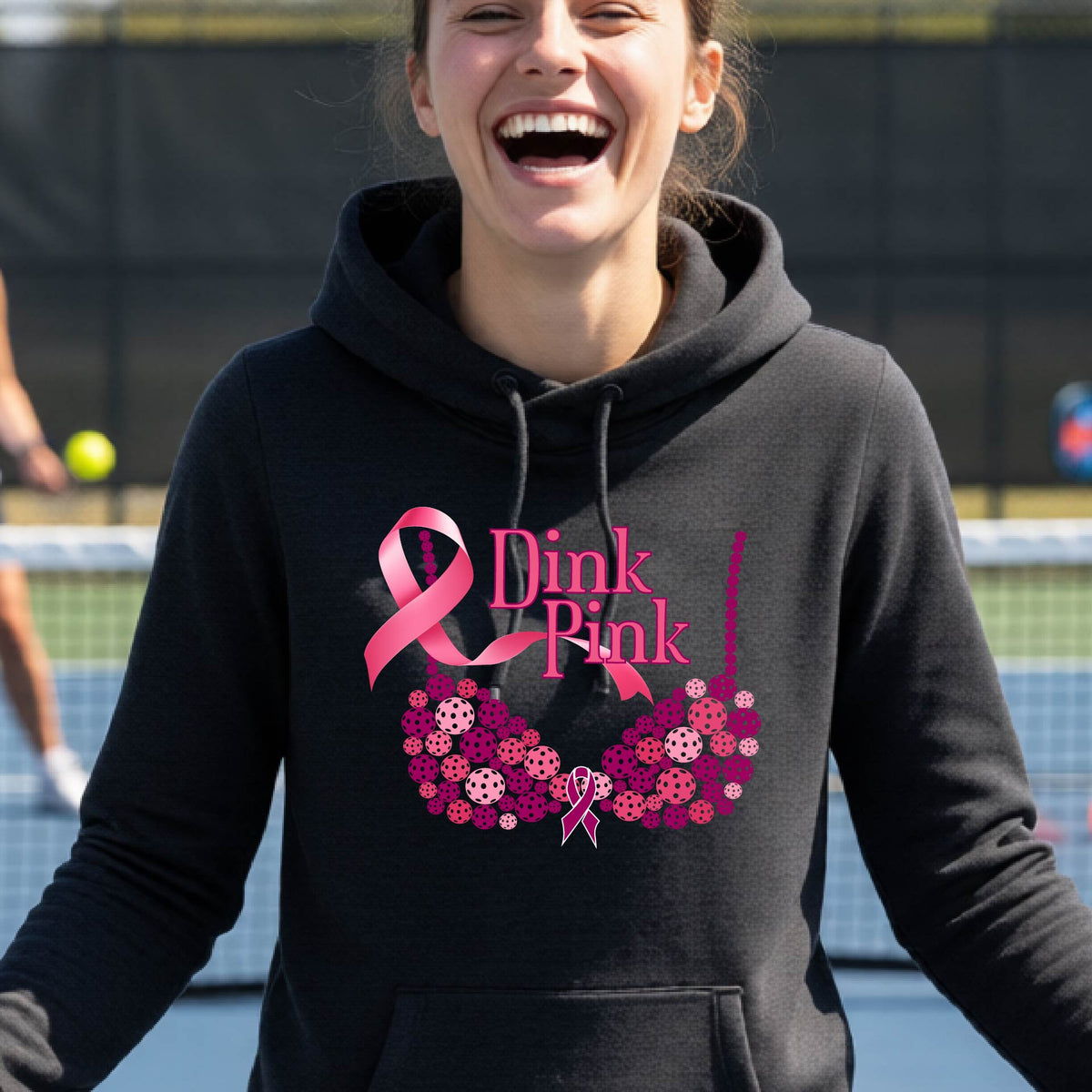 A woman smiling on a pickleball court wearing a black hoodie with a pink 'Dink Pink' graphic made of pickleball and a ribbon and a bra-shaped floral design.