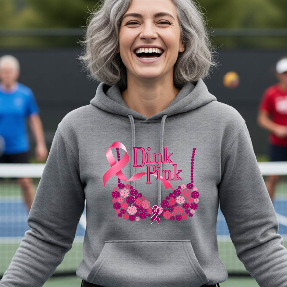 A woman smiling on a pickleball court wearing a gray hoodie with a pink 'Dink Pink' graphic made of pickleball and a ribbon and a bra-shaped floral design.