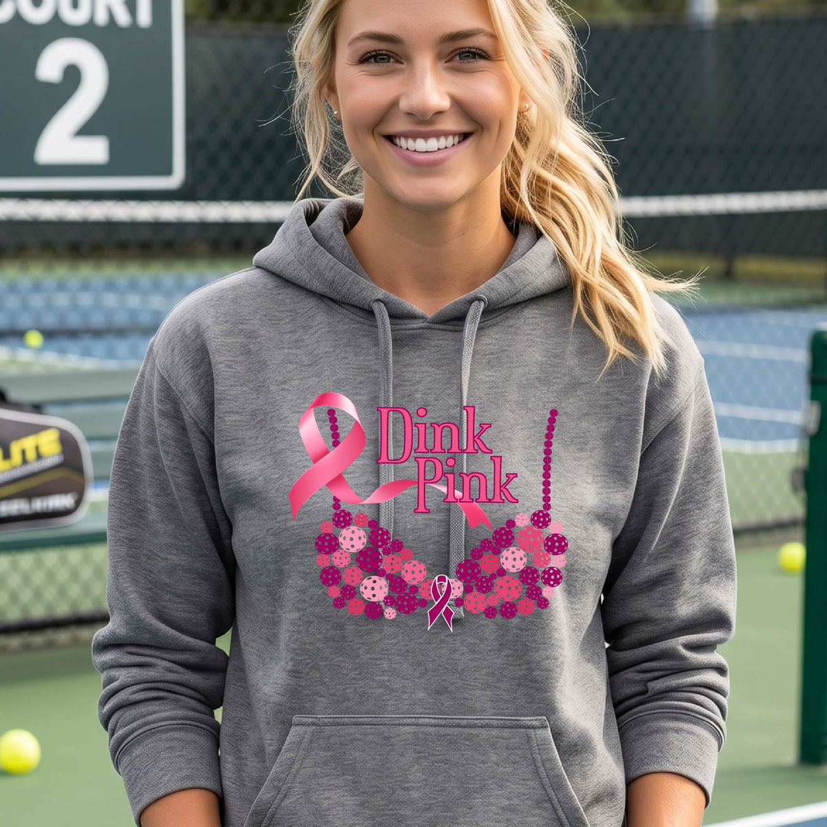 A woman smiling on a pickleball court wearing a gray hoodie with a pink 'Dink Pink' graphic made of pickleball and a ribbon and a bra-shaped floral design.