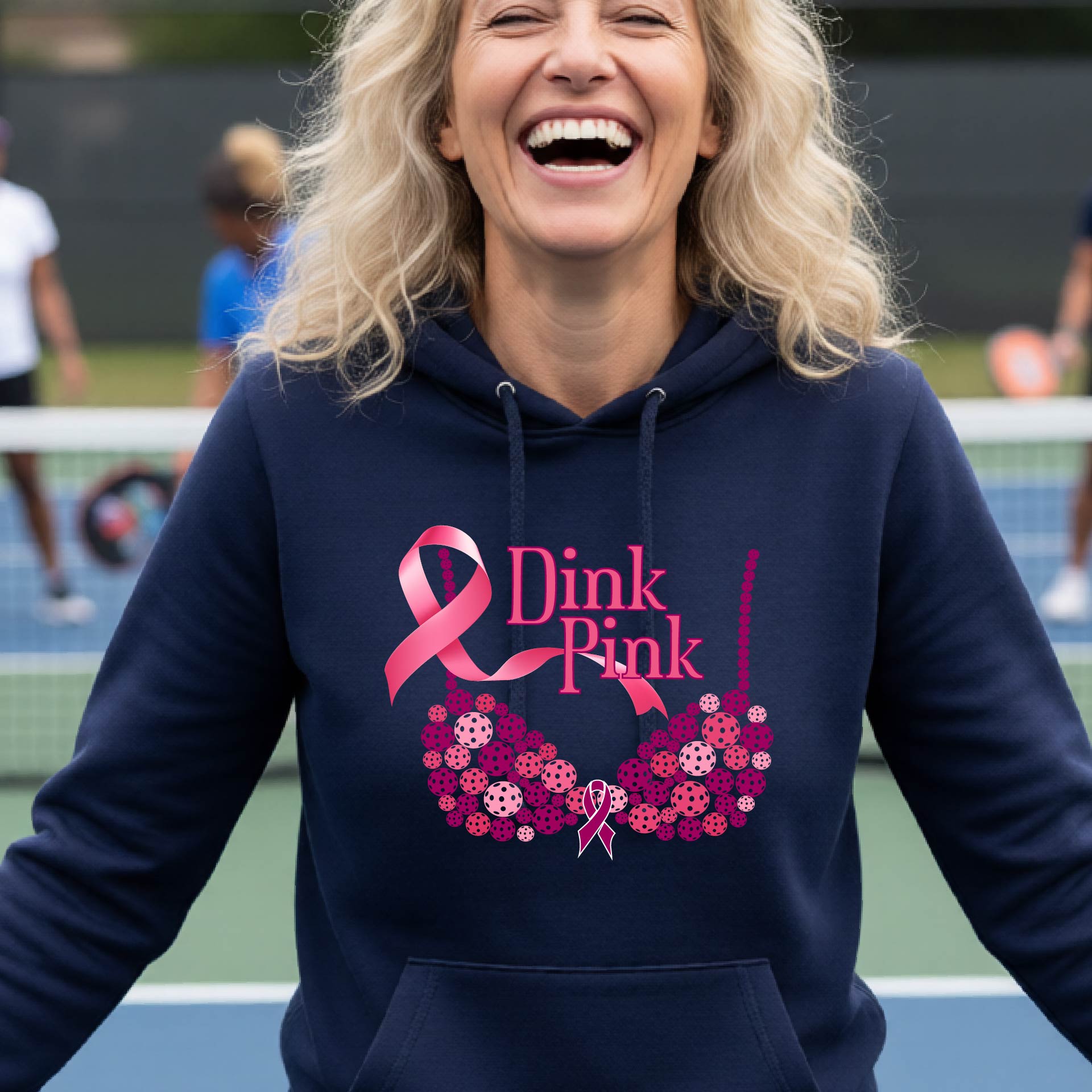 A woman smiling on a pickleball court wearing a navy blue hoodie with a pink 'Dink Pink' graphic made of pickleball and a ribbon and a bra-shaped floral design.