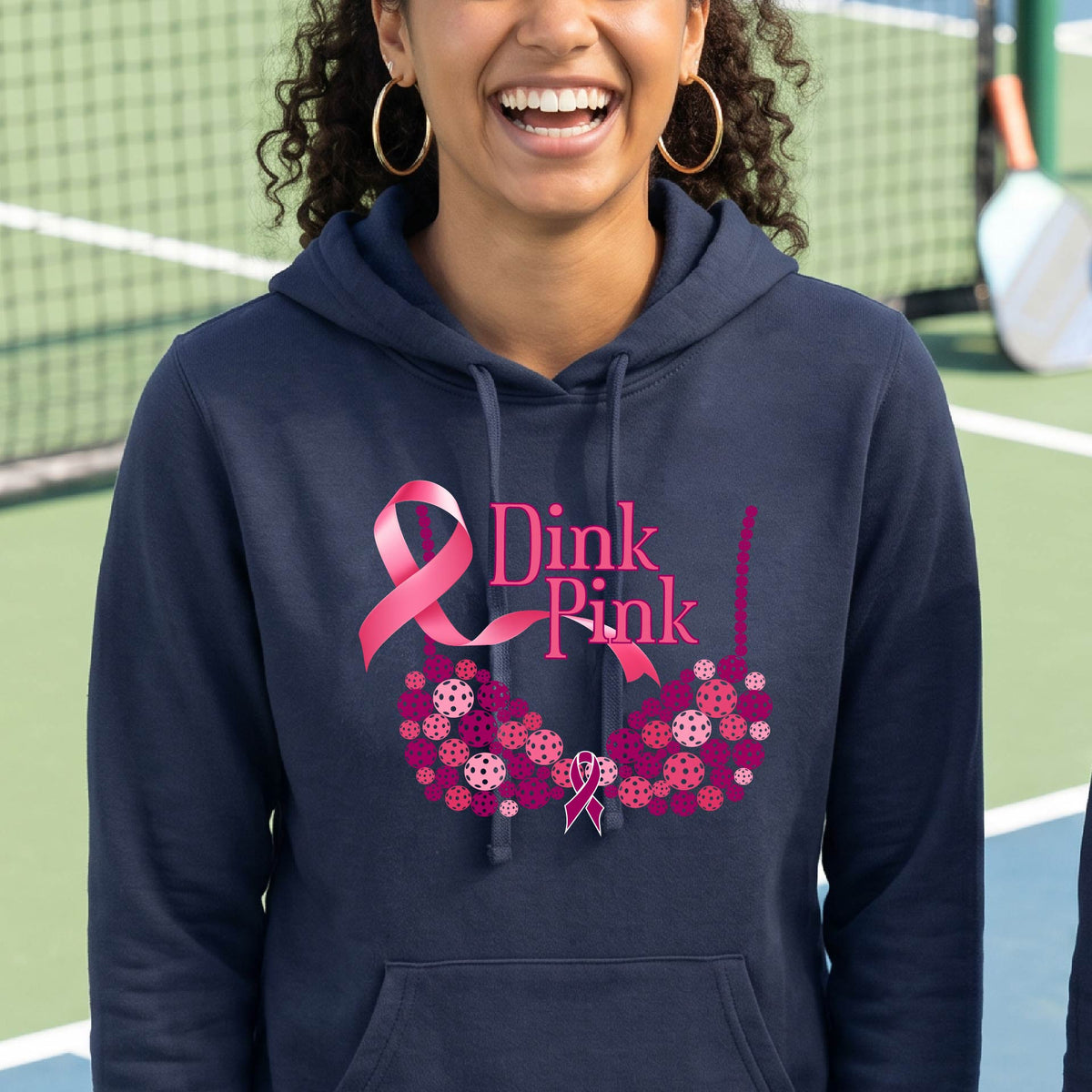 A woman smiling on a pickleball court wearing a navy blue hoodie with a pink 'Dink Pink' graphic made of pickleball and a ribbon and a bra-shaped floral design.