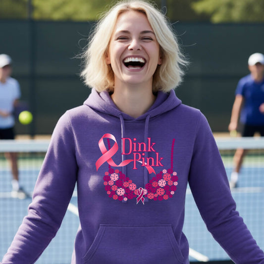A woman smiling on a pickleball court wearing a purple hoodie with a pink 'Dink Pink' graphic made of pickleball and a ribbon and a bra-shaped floral design.
