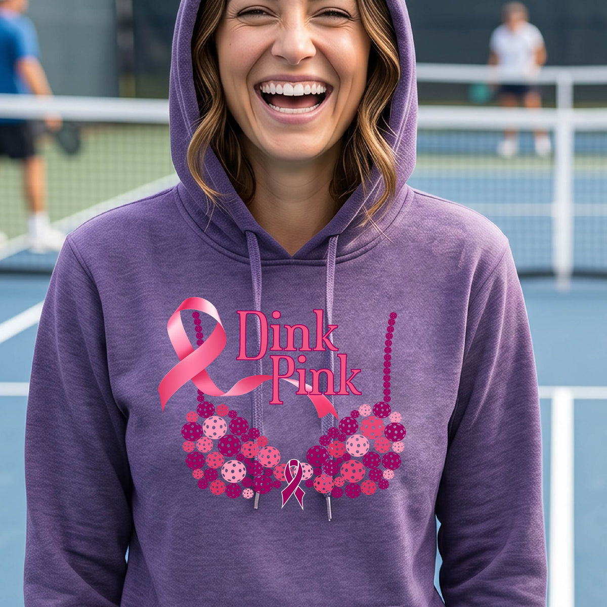 A woman smiling on a pickleball court wearing a purple hoodie with a pink 'Dink Pink' graphic made of pickleball and a ribbon and a bra-shaped floral design.