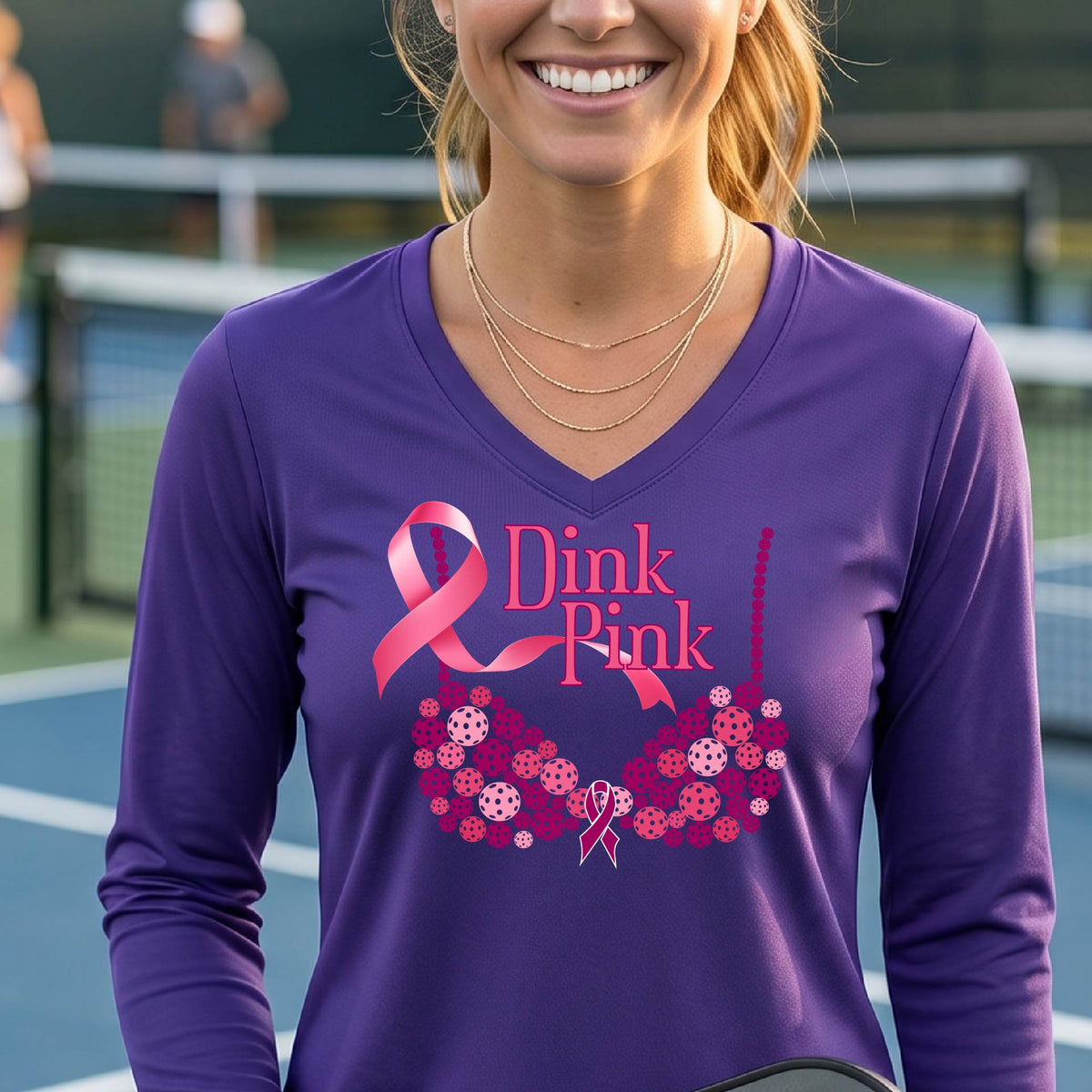 A woman on a pickleball court wearing a purple long-sleeve V-neck shirt with a pink 'Dink Pink' breast cancer awareness graphic and floral bra design made of pickleballs.