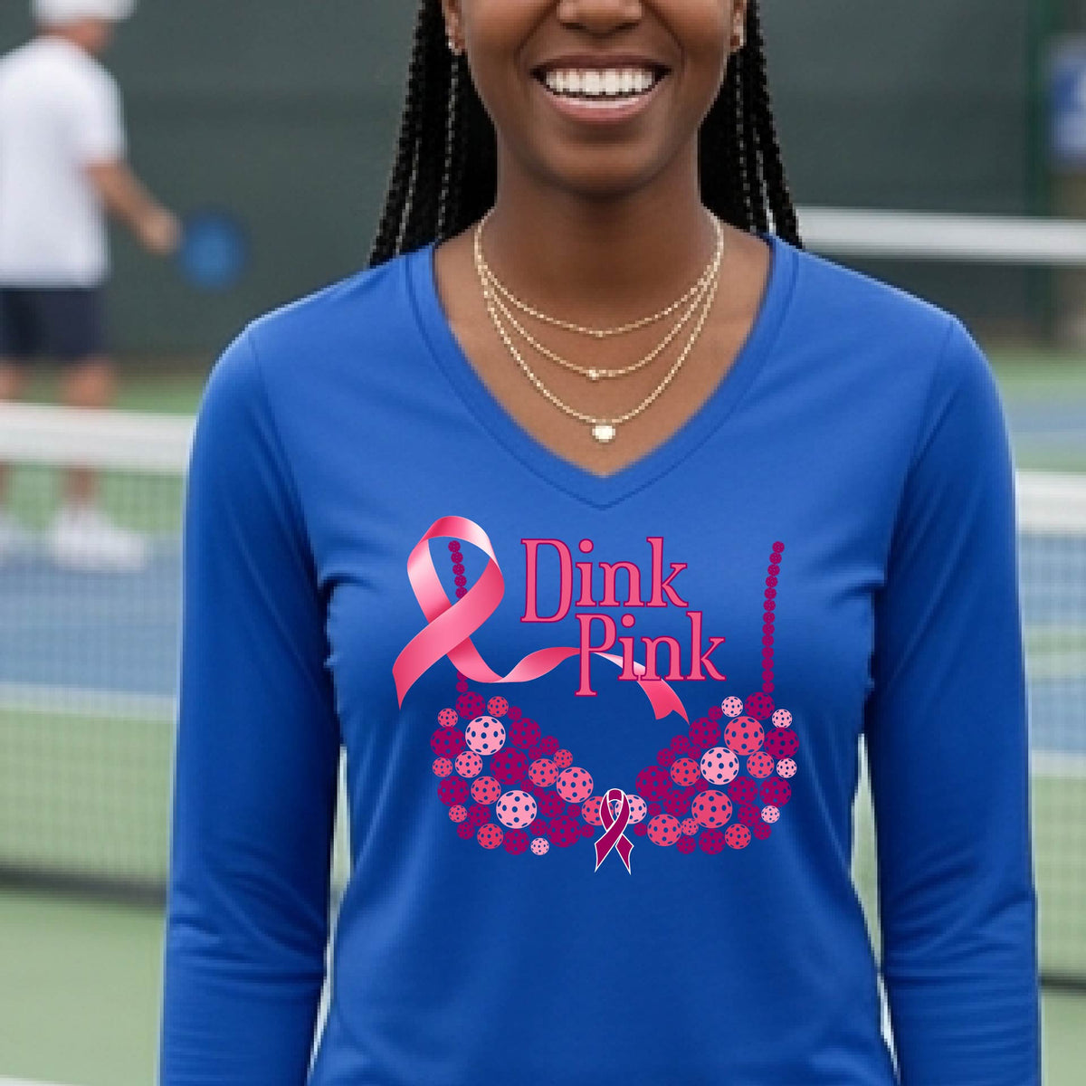 A woman on a pickleball court wearing a royal blue long-sleeve V-neck shirt with a pink 'Dink Pink' breast cancer awareness graphic and floral bra design made of pickleballs.