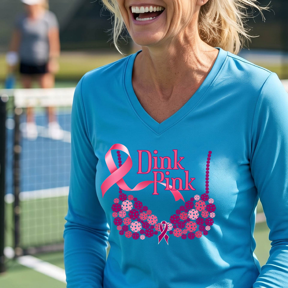 A woman on a pickleball court wearing a bright blue long-sleeve V-neck shirt with a pink 'Dink Pink' breast cancer awareness graphic and floral bra design made of pickleballs.