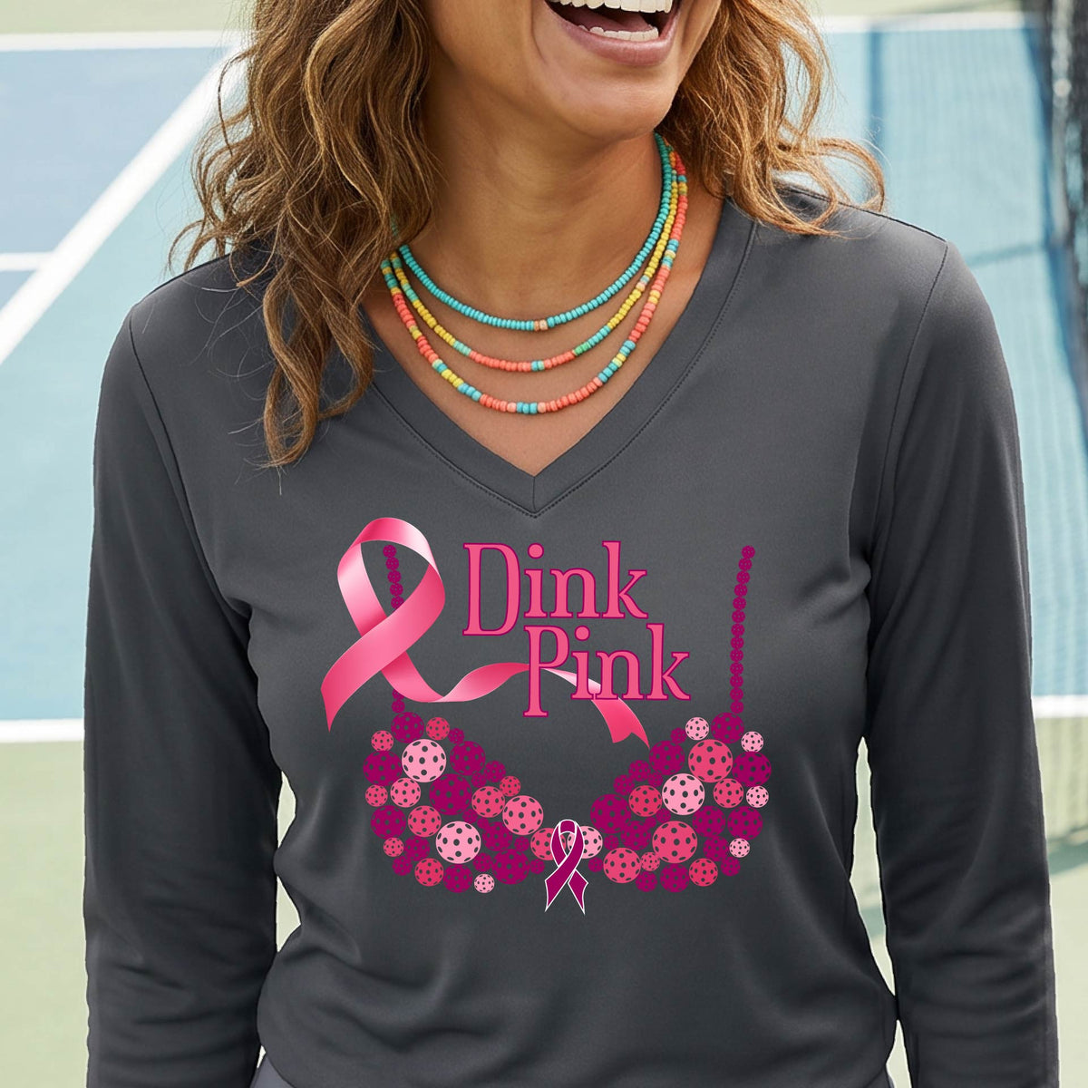 A woman on a pickleball court wearing a gray long-sleeve V-neck shirt with a pink 'Dink Pink' breast cancer awareness graphic and floral bra design made of pickleballs.