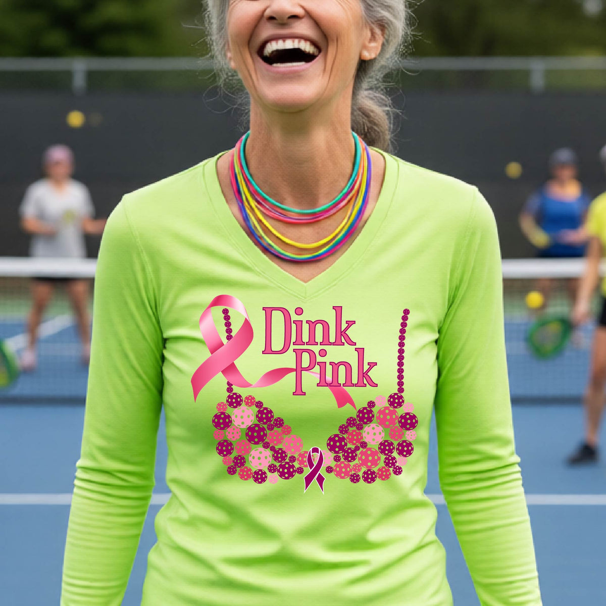 A woman on a pickleball court wearing a bright lime green long-sleeve V-neck shirt with a pink 'Dink Pink' breast cancer awareness graphic and floral bra design made of pickleballs.