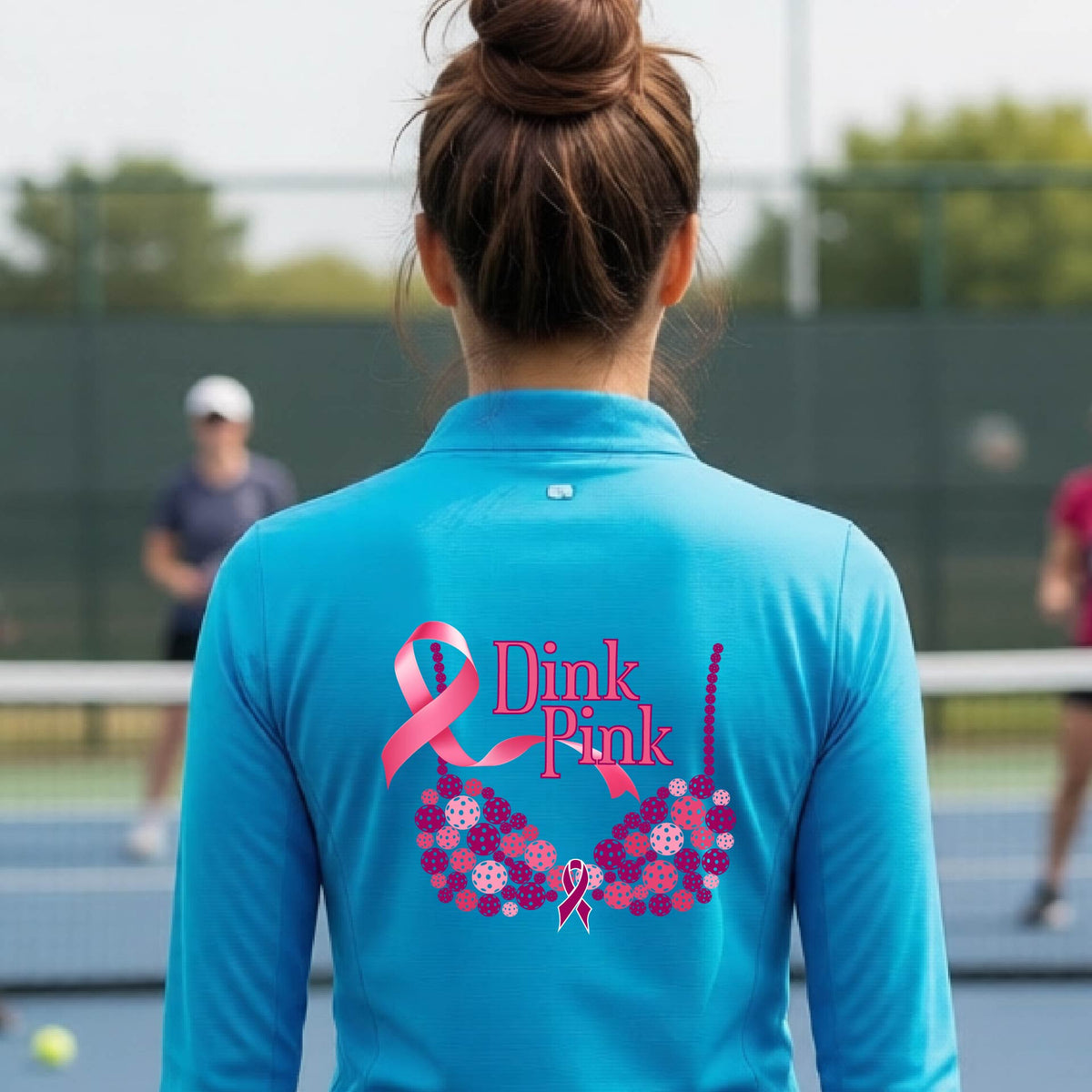 A woman viewed from behind on a pickleball court, wearing a bright blue long-sleeve athletic quarter-zip shirt with a large pink and purple 'Dink Pink' bra graphic made of pickleballs and ribbon design printed on the back.