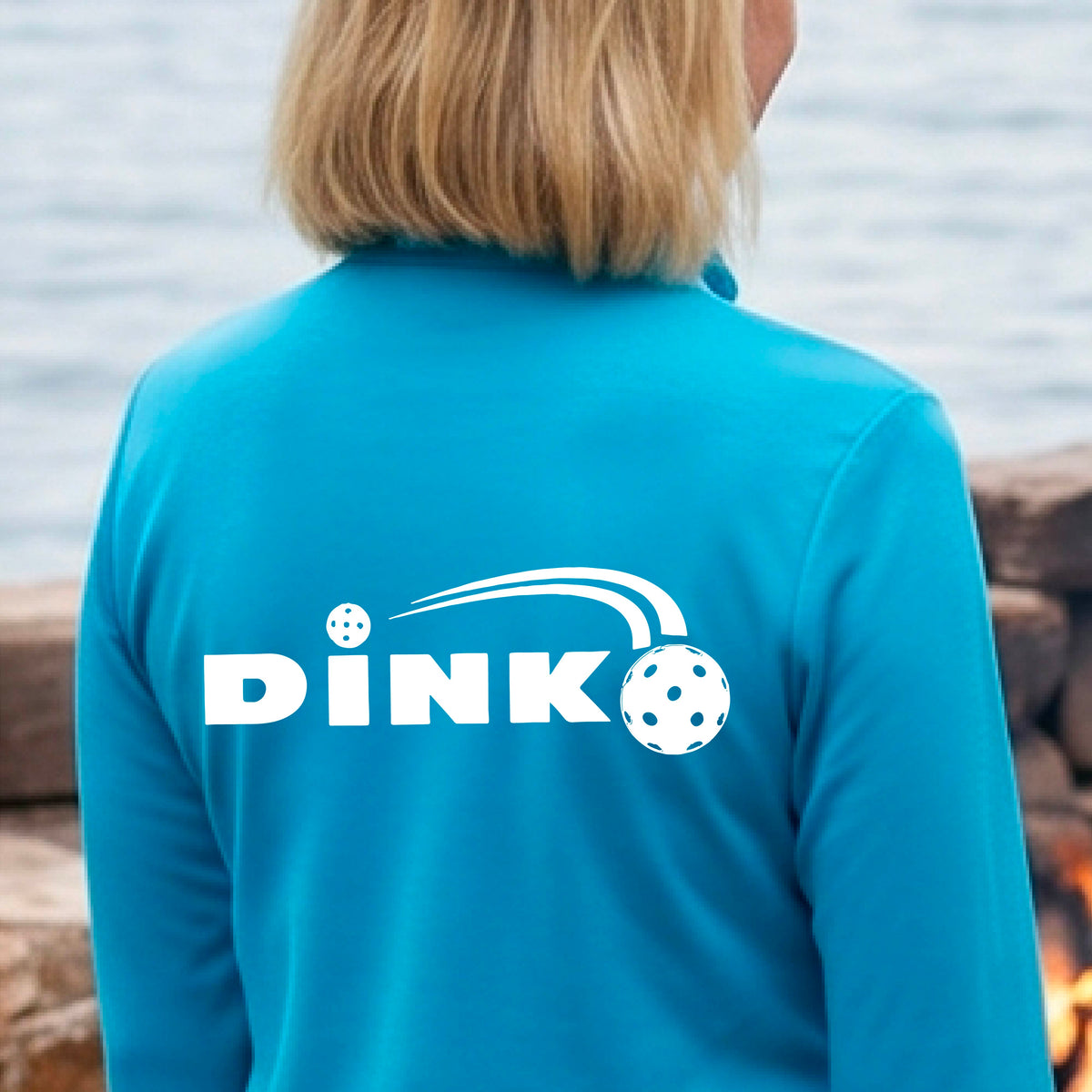 A woman on a pickleball court wearing a bright blue long-sleeve quarter-zip jacket with a white 'DINK' logo and pickleball in flight printed across the back.