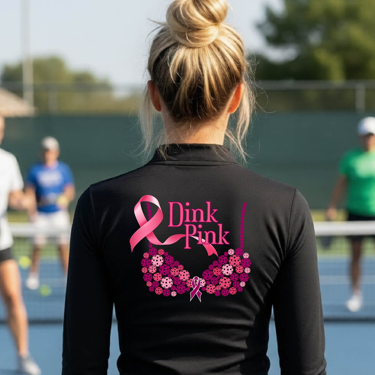 A woman viewed from behind on a pickleball court, wearing a black long-sleeve athletic quarter-zip shirt with a large pink and purple 'Dink Pink' bra graphic made of pickleballs and ribbon design printed on the back.