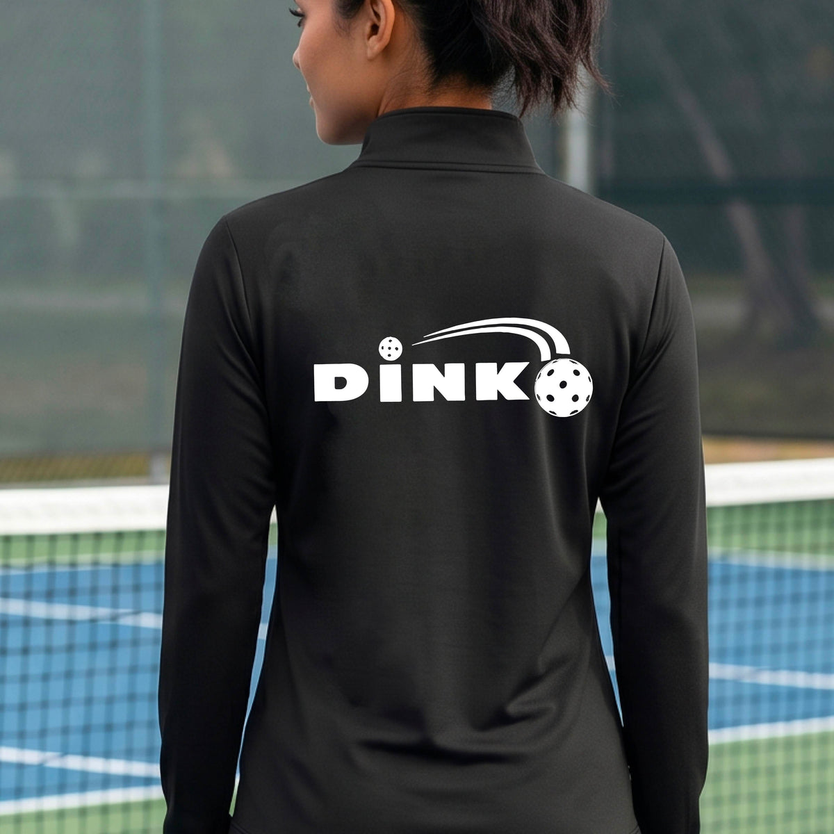 A woman on a pickleball court wearing a black long-sleeve quarter-zip jacket with a white 'DINK' logo and pickleball in flight printed across the back.