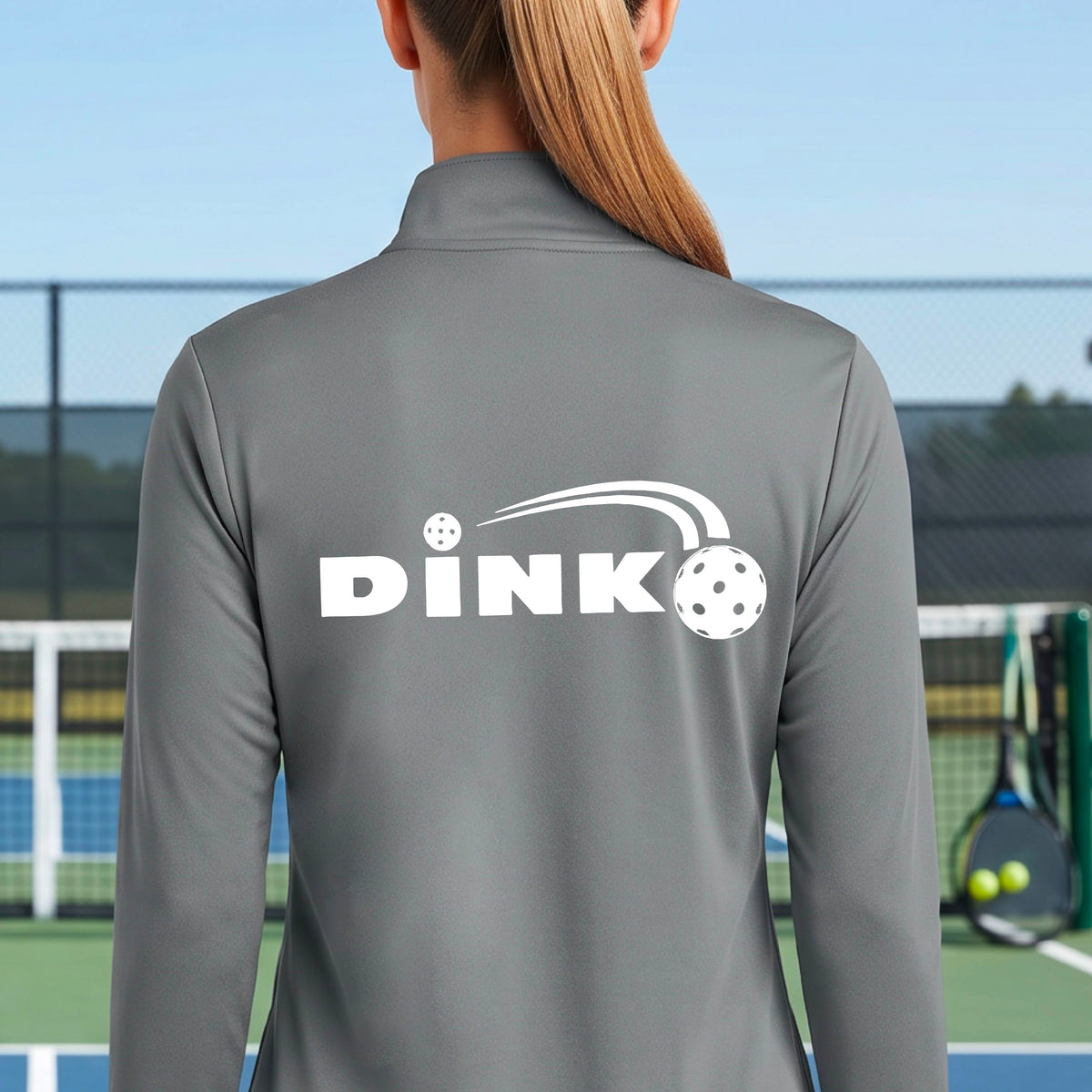 A woman on a pickleball court wearing a gray long-sleeve quarter-zip jacket with a white 'DINK' logo and pickleball in flight printed across the back.