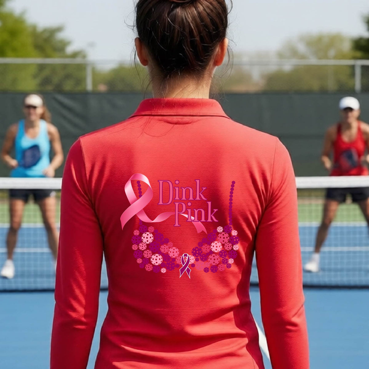 A woman viewed from behind on a pickleball court, wearing a red long-sleeve athletic quarter-zip shirt with a large pink and purple 'Dink Pink' bra graphic made of pickleballs and ribbon design printed on the back.