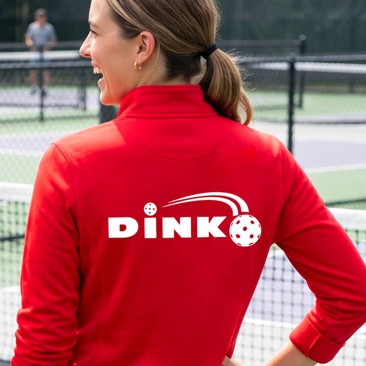 A woman on a pickleball court wearing a bright red long-sleeve quarter-zip jacket with a white 'DINK' logo and pickleball in flight printed across the back.