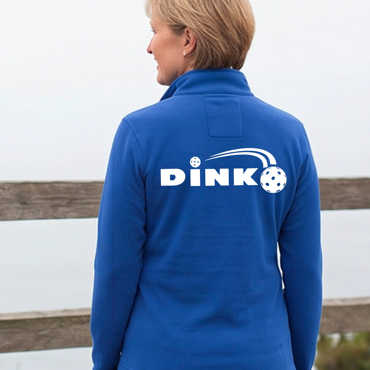 A woman on a pickleball court wearing a bright royal blue long-sleeve quarter-zip jacket with a white 'DINK' logo and pickleball in flight printed across the back.