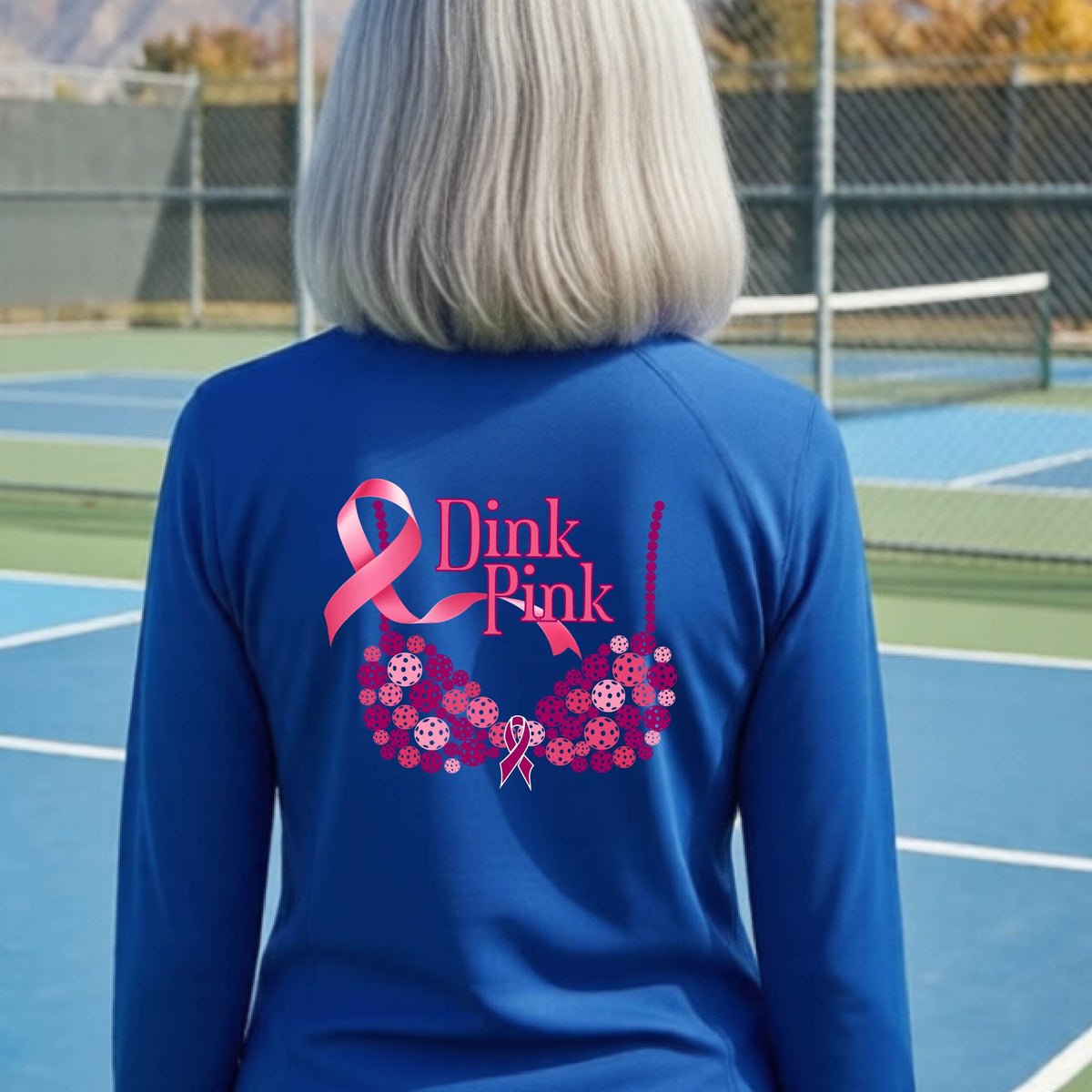 A woman viewed from behind on a pickleball court, wearing a bright royal blue long-sleeve athletic quarter-zip shirt with a large pink and purple 'Dink Pink' bra graphic made of pickleballs and ribbon design printed on the back.