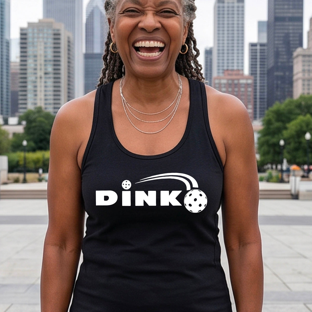 A close up of a black racerback tank top with a white design "DINK" and a white pickleball in flight over the words, on the front of the shirt, standing on a pickleball court. 