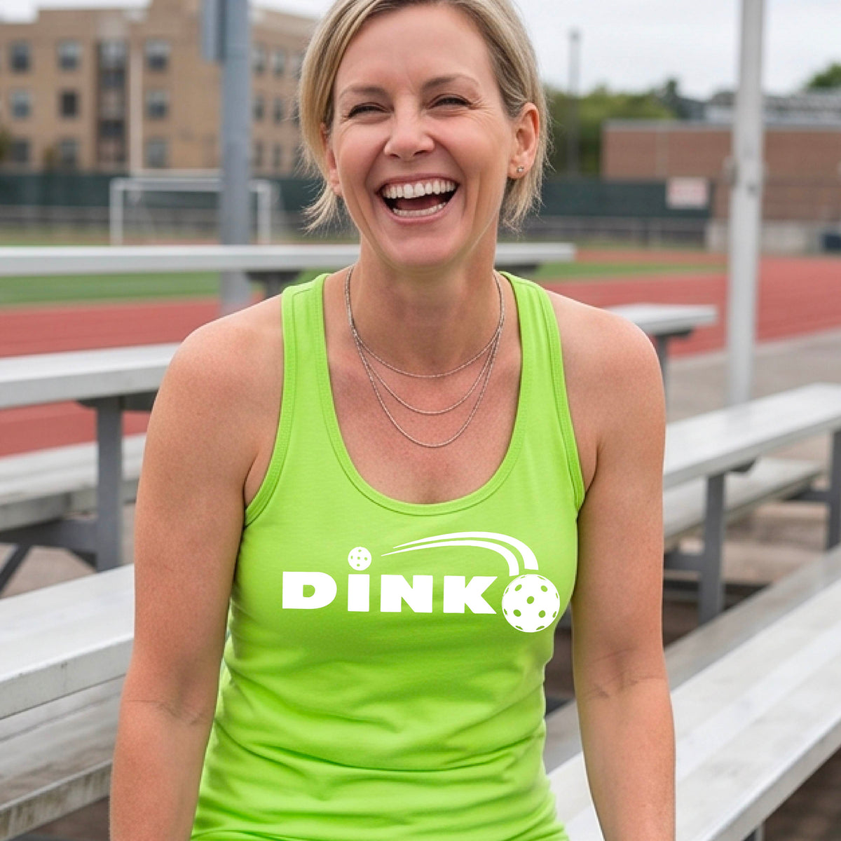 A close up of a lime green racerback tank top with a white design "DINK" and a white pickleball in flight over the words, on the front of the shirt, standing on a pickleball court. 