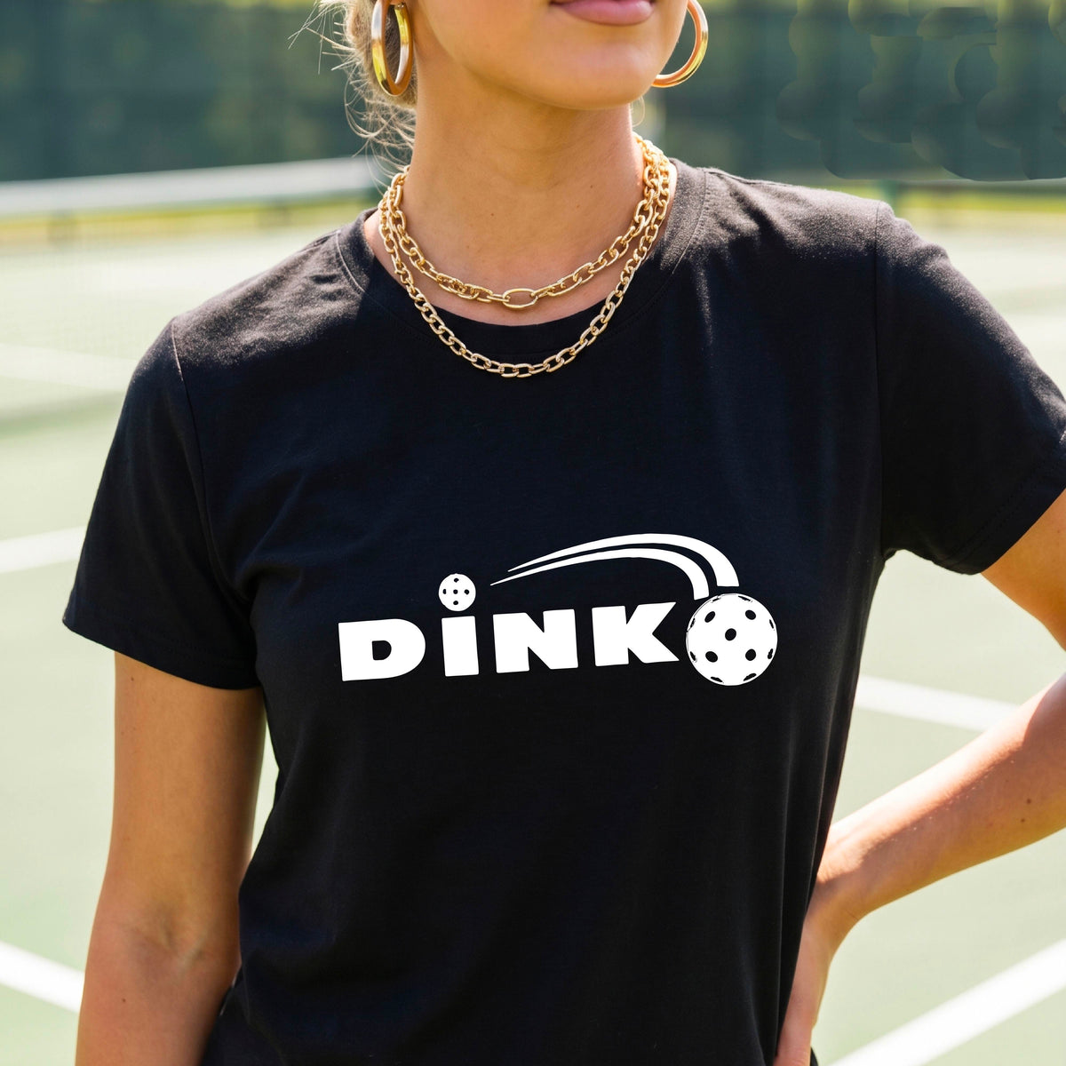A woman wearing a black short-sleeve v-neck t-shirt with a pickleball design of text 'DINK' with a white pickleall in flight, standing on a pickleball court.