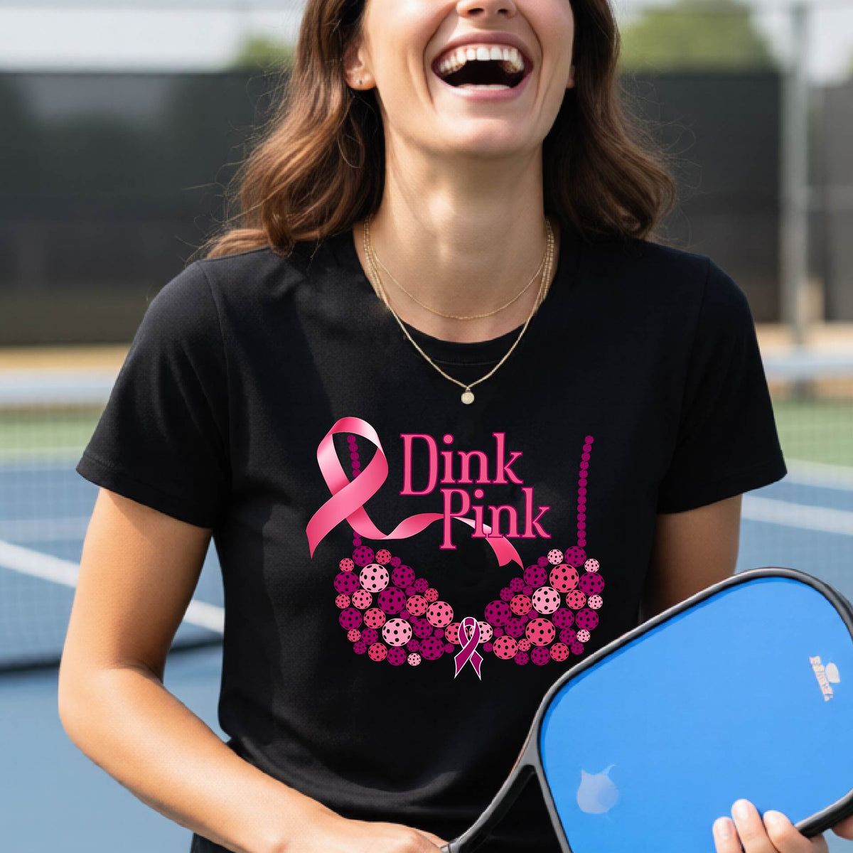 A woman wearing a black graphic t-shirt with a pink and purple bra-shaped design and the words 'Dink Pink' on the front, set against a blurred pickleball court background.