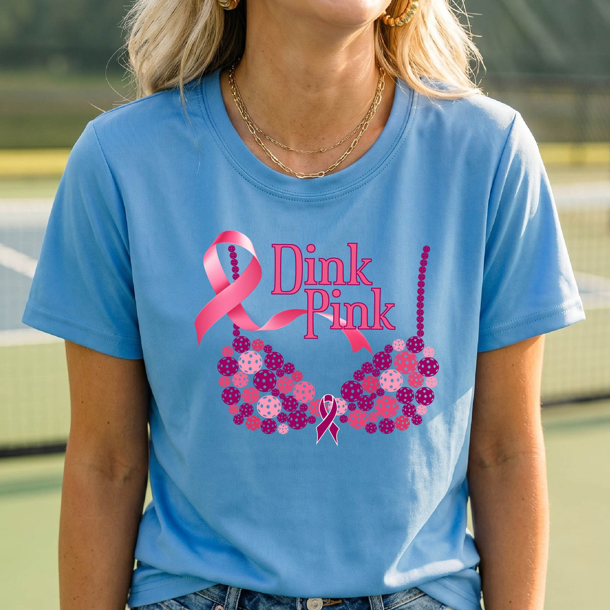 A woman wearing a light blue graphic t-shirt with a pink and purple bra-shaped design and the words 'Dink Pink' on the front, set against a blurred pickleball court background.