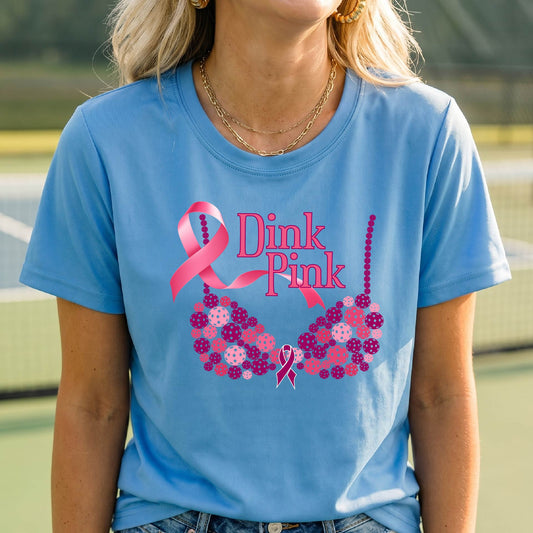 A woman wearing a light blue graphic t-shirt with a pink and purple bra-shaped design and the words 'Dink Pink' on the front, set against a blurred pickleball court background.