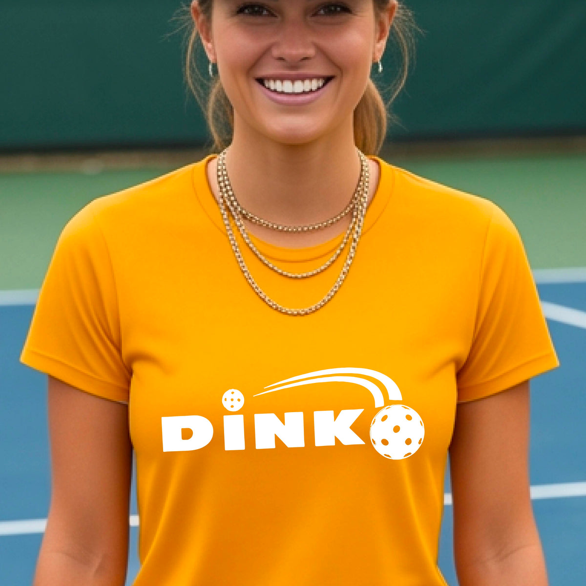 A woman wearing a mustard yellow gold short-sleeve v-neck t-shirt with a pickleball design of text 'DINK' with a white pickleall in flight, standing on a pickleball court.