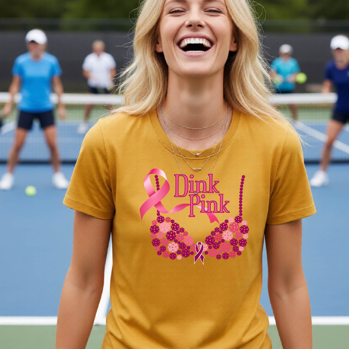 A woman wearing a mustard yellow gold graphic t-shirt with a pink and purple bra-shaped design and the words 'Dink Pink' on the front, set against a blurred pickleball court background.