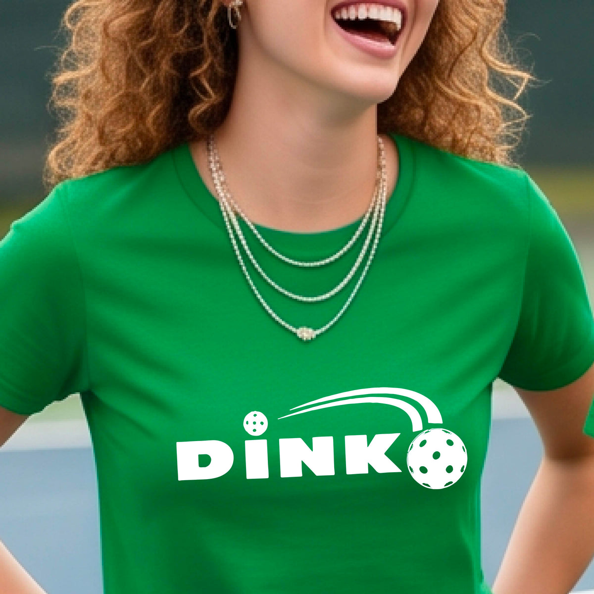 A woman wearing a green short-sleeve v-neck t-shirt with a pickleball design of text 'DINK' with a white pickleall in flight, standing on a pickleball court.