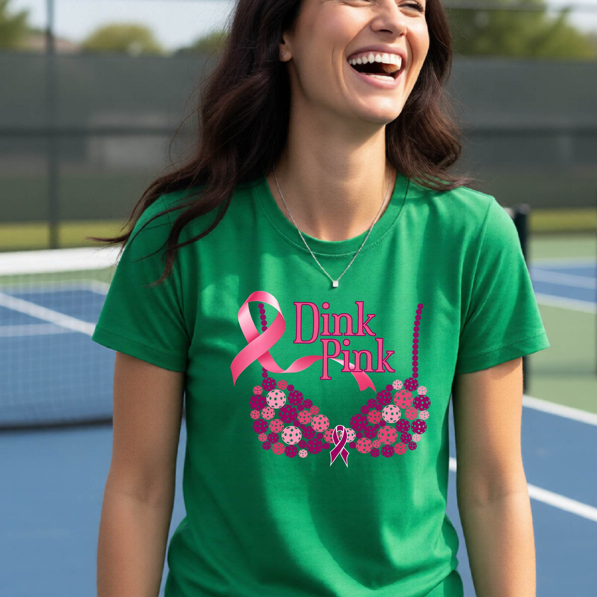 A woman wearing a green graphic t-shirt with a pink and purple bra-shaped design and the words 'Dink Pink' on the front, set against a blurred pickleball court background.