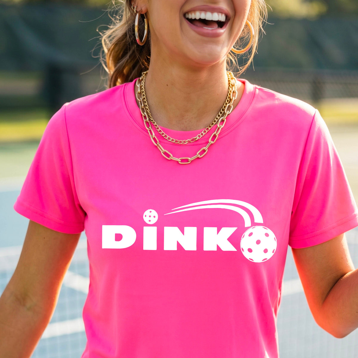 A woman wearing a pink short-sleeve v-neck t-shirt with a pickleball design of text 'DINK' with a white pickleall in flight, standing on a pickleball court.