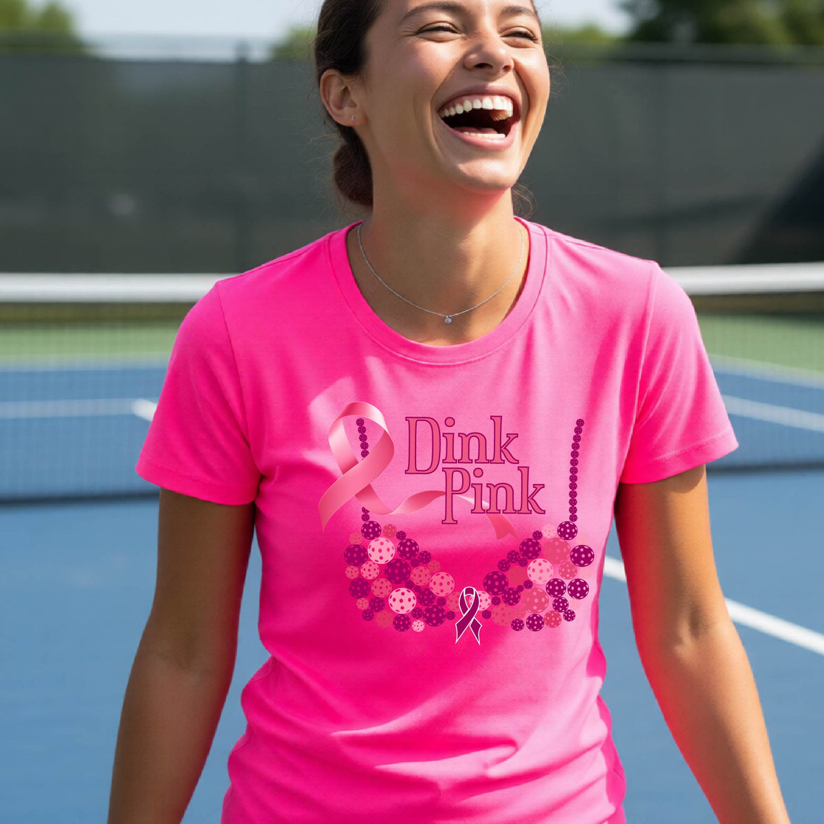 A woman wearing a pink graphic t-shirt with a pink and purple bra-shaped design and the words 'Dink Pink' on the front, set against a blurred pickleball court background.