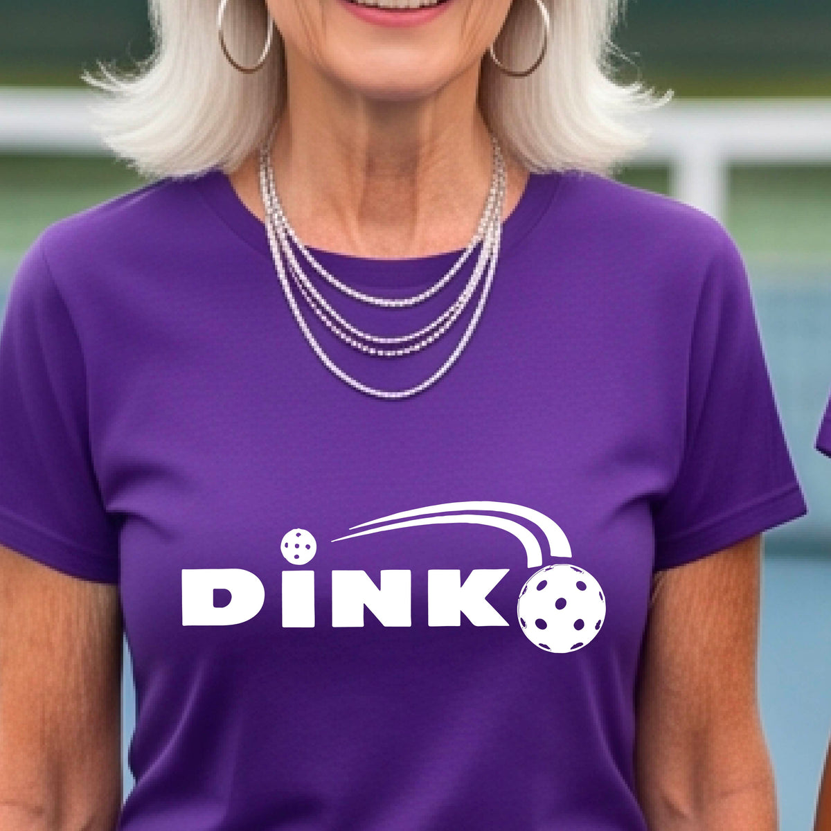 A woman wearing a purple short-sleeve v-neck t-shirt with a pickleball design of text 'DINK' with a white pickleall in flight, standing on a pickleball court.