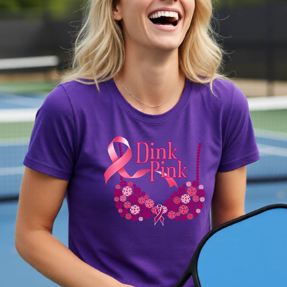 A woman wearing a purple graphic t-shirt with a pink and purple bra-shaped design and the words 'Dink Pink' on the front, set against a blurred pickleball court background.
