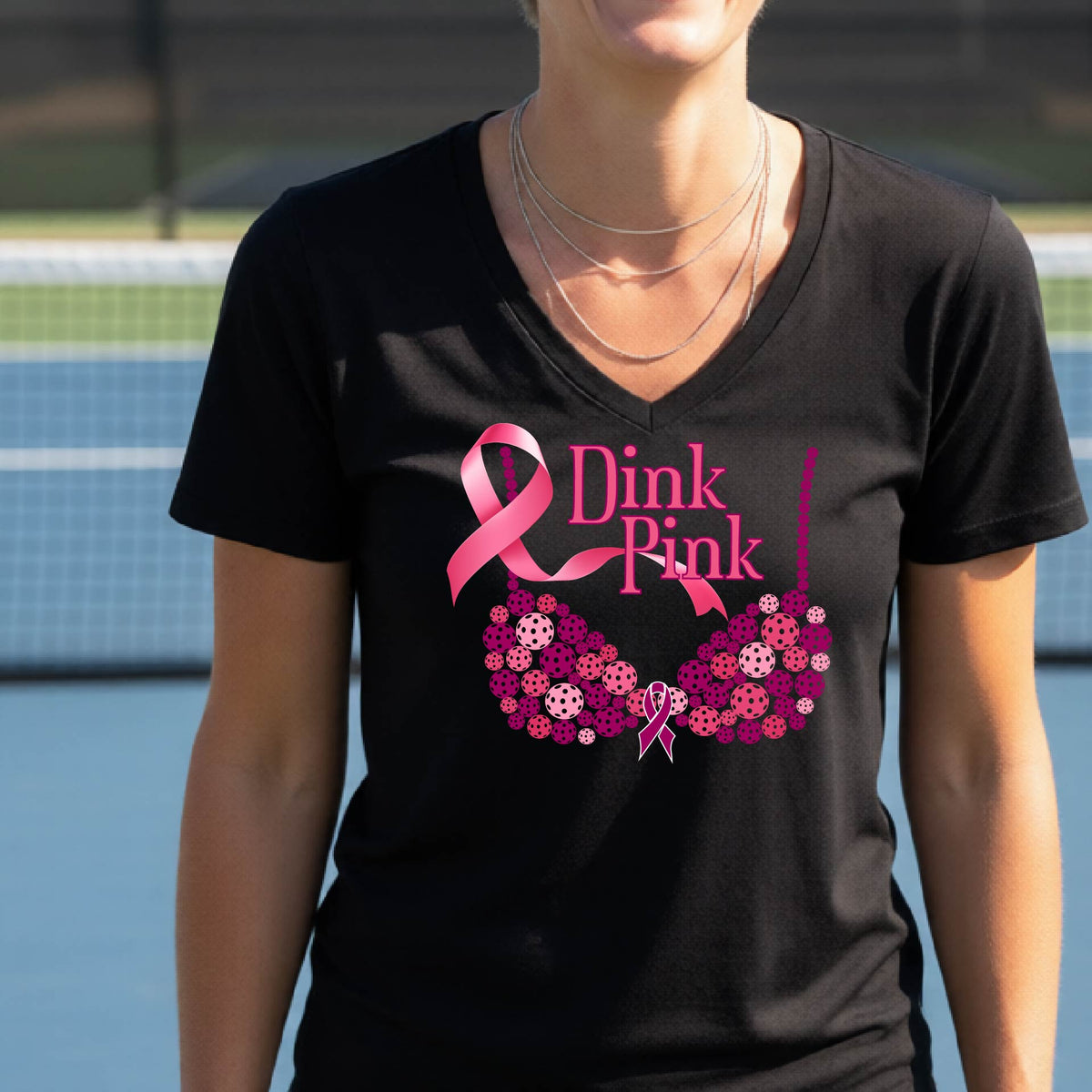 A woman wearing a black vneck t-shirt "Dink Pink" ' breast cancer awareness graphic on the front, set against a blurred pickleball court background.