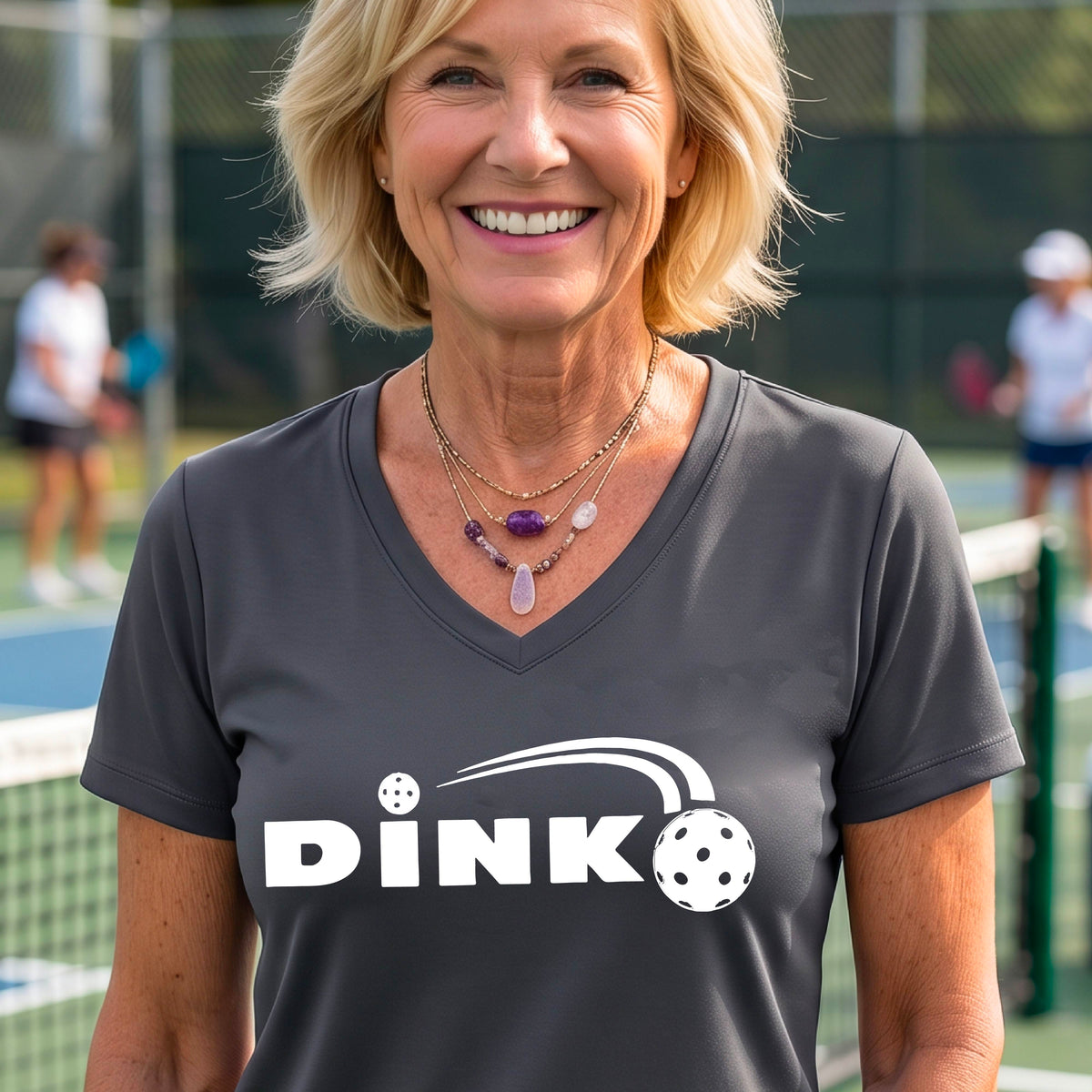 A woman wearing a gray short-sleeve v-neck t-shirt with a pickleball design of text 'DINK' with a white pickleall in flight, standing on a pickleball court.