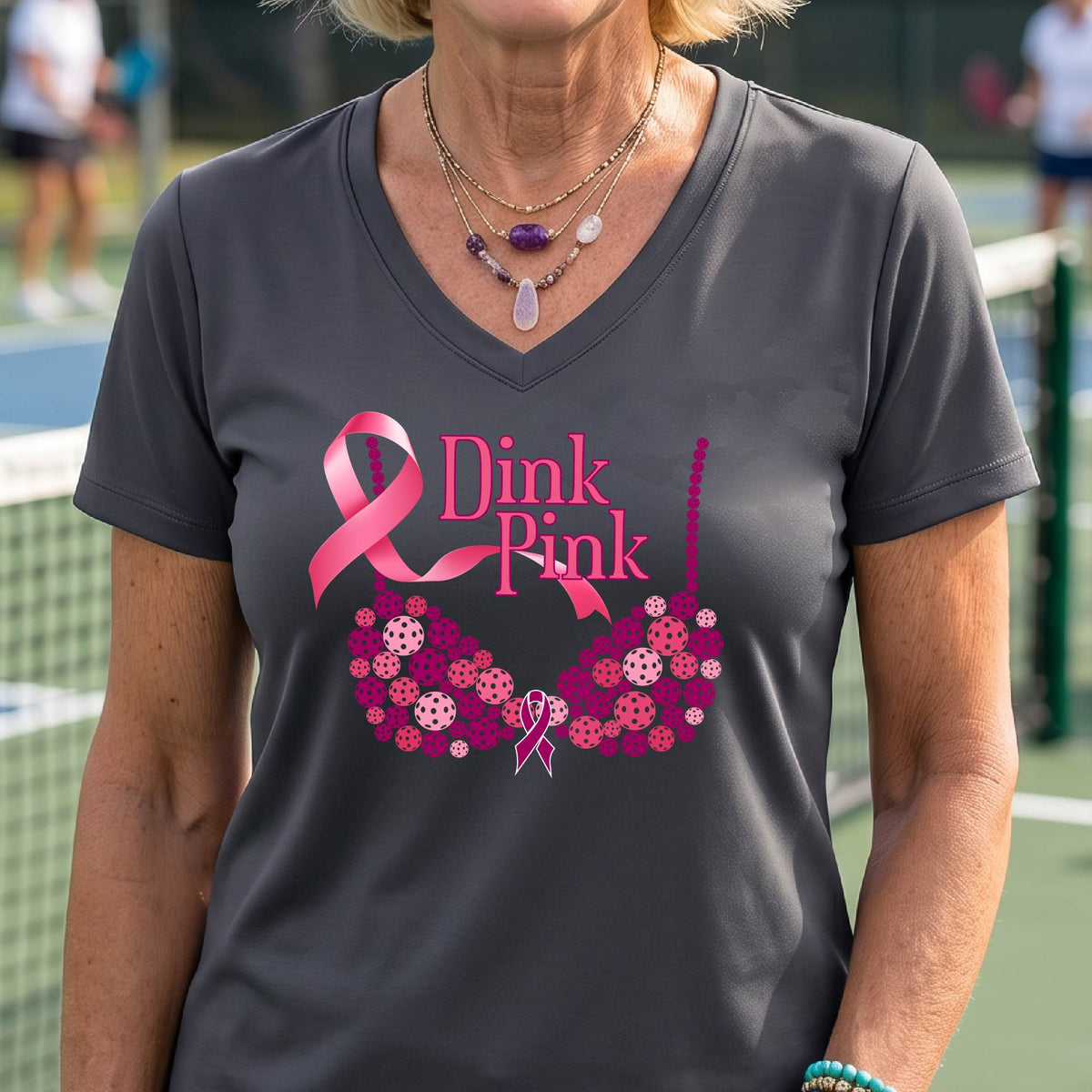 A woman wearing a dark gray V-neck t-shirt with a pink 'Dink Pink' breast cancer awareness graphic on the front, set against a blurred pickleball court background.