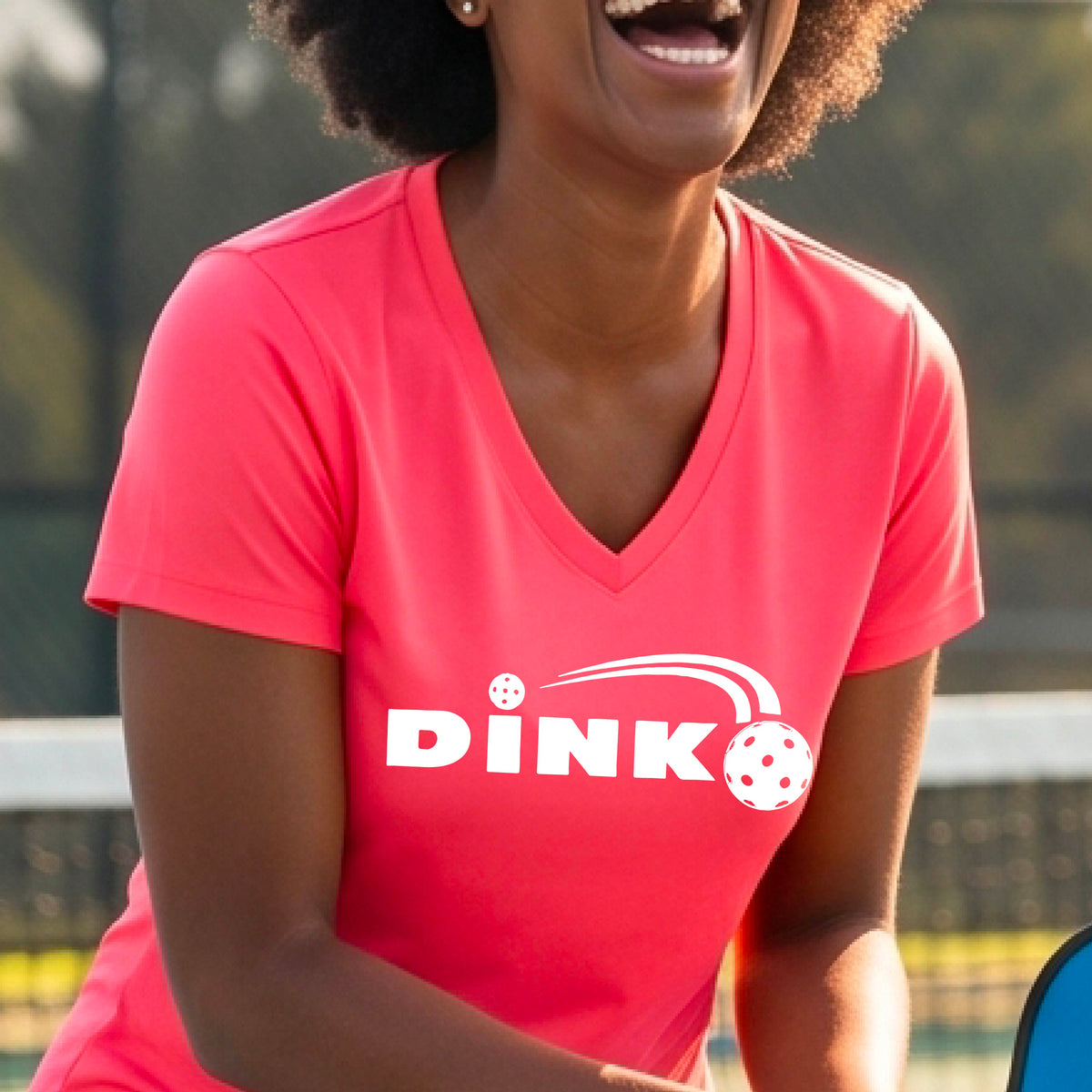 A woman wearing a coral pink short-sleeve v-neck t-shirt with a pickleball design of text 'DINK' with a white pickleall in flight, standing on a pickleball court.