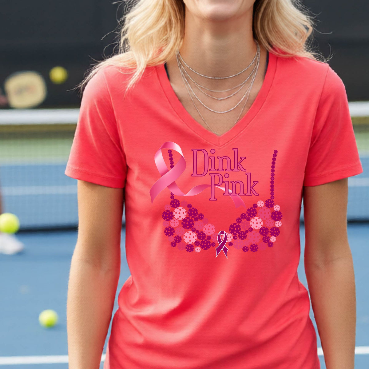 A woman wearing a coral pink V-neck t-shirt with a pink 'Dink Pink' breast cancer awareness graphic on the front, set against a blurred pickleball court background.