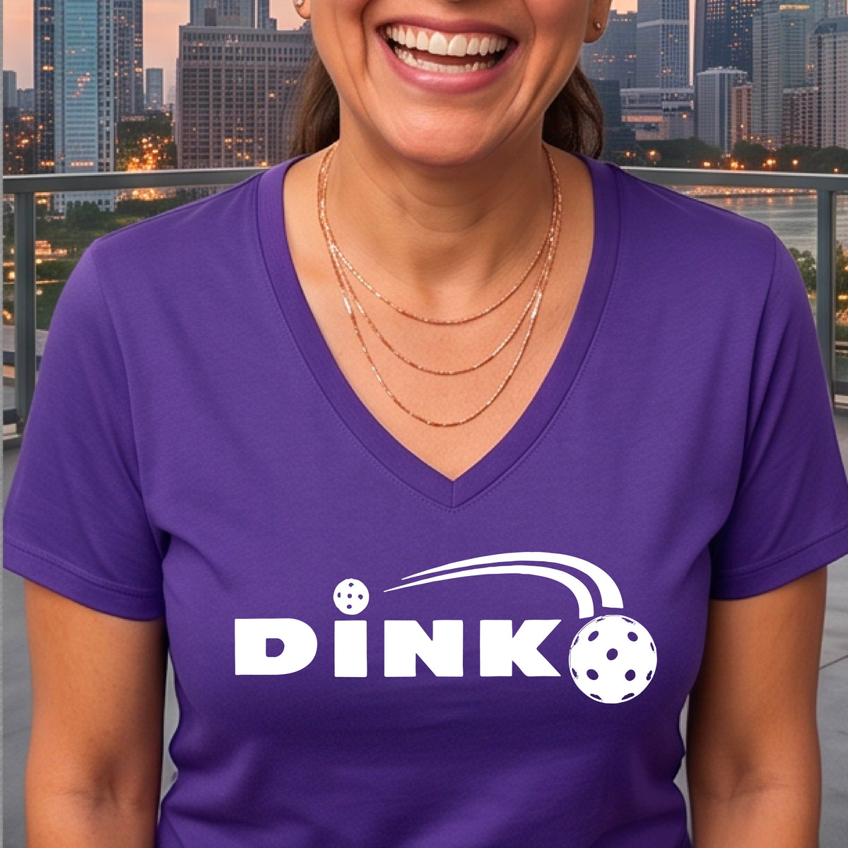 A woman wearing a purple short-sleeve v-neck t-shirt with a pickleball design of text 'DINK' with a white pickleall in flight, standing on a pickleball court.