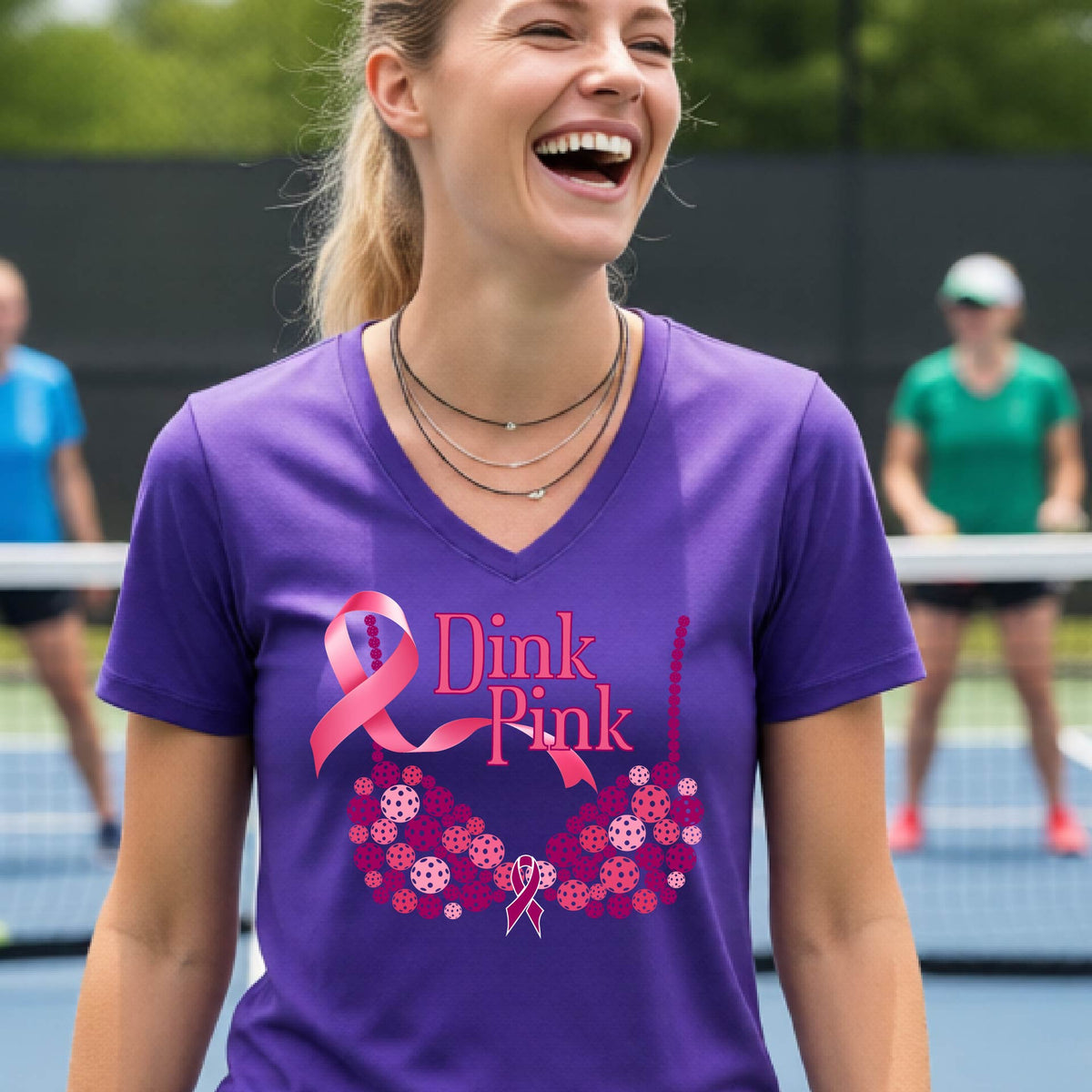 A woman wearing a purple V-neck t-shirt with a pink 'Dink Pink' breast cancer awareness graphic on the front, set against a blurred pickleball court background.