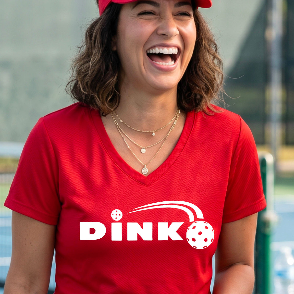 A woman wearing a red short-sleeve v-neck t-shirt with a pickleball design of text 'DINK' with a white pickleall in flight, standing on a pickleball court.