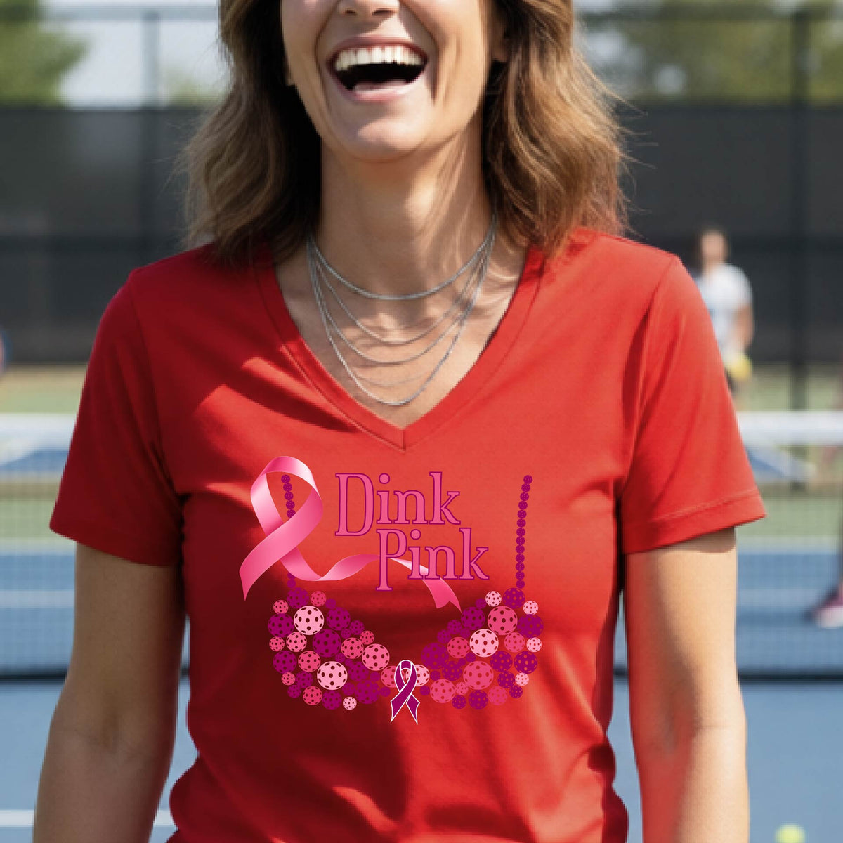 A woman wearing a red V-neck t-shirt with a pink 'Dink Pink' breast cancer awareness graphic on the front, set against a blurred pickleball court background.