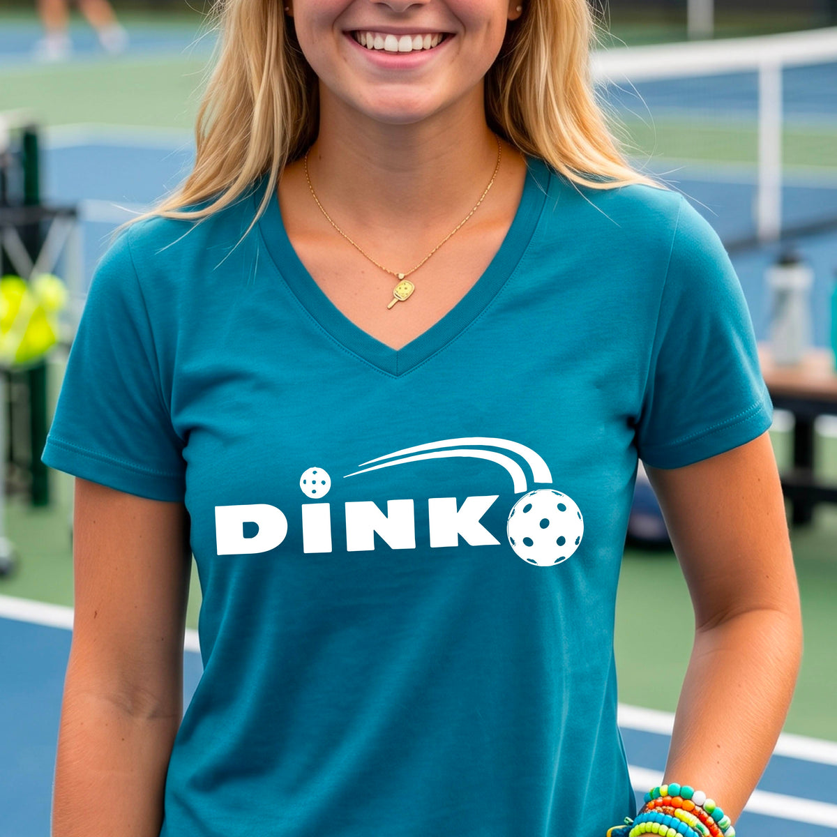 A woman wearing a teal short-sleeve v-neck t-shirt with a pickleball design of text 'DINK' with a white pickleall in flight, standing on a pickleball court.