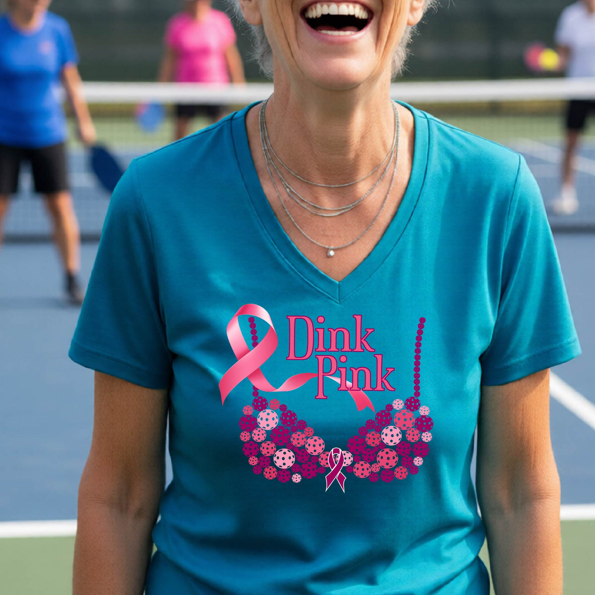 A woman wearing a teal V-neck t-shirt with a pink 'Dink Pink' breast cancer awareness graphic on the front, set against a blurred pickleball court background.