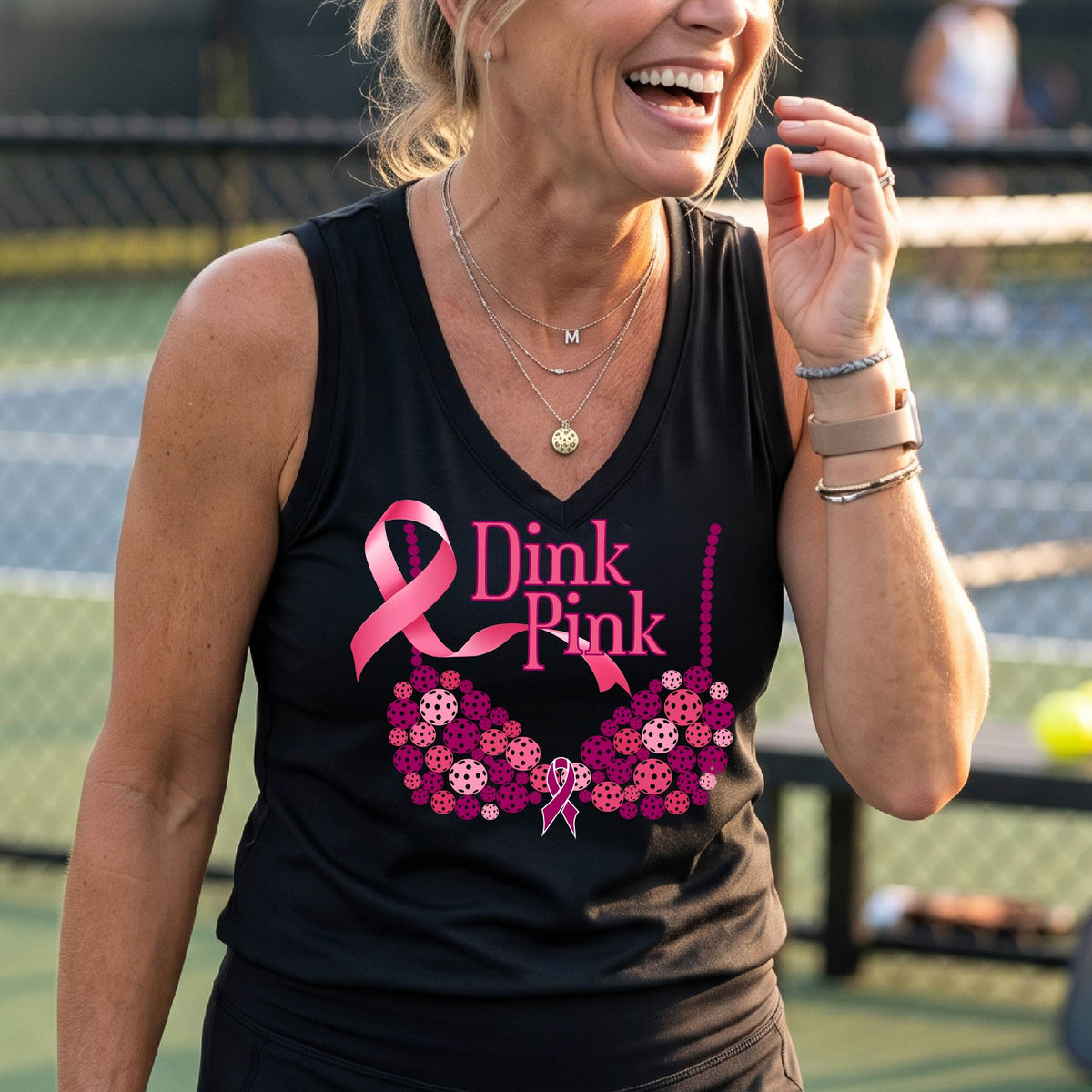 A woman on a pickleball court wearing a black sleeveless  top with a pink 'Dink Pink' graphic and a pink floral bra design. The background shows a blurred pickleball court and net.