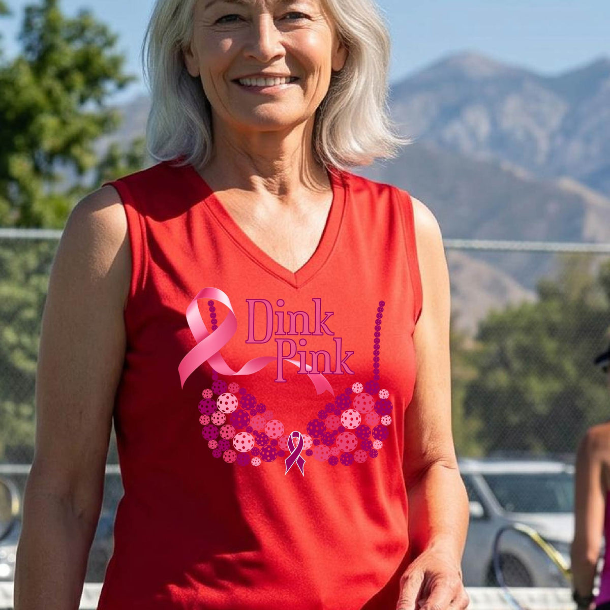 A woman on a pickleball court wearing a red sleeveless top with a pink 'Dink Pink' graphic and a pink floral bra design. The background shows a blurred pickleball court and net.