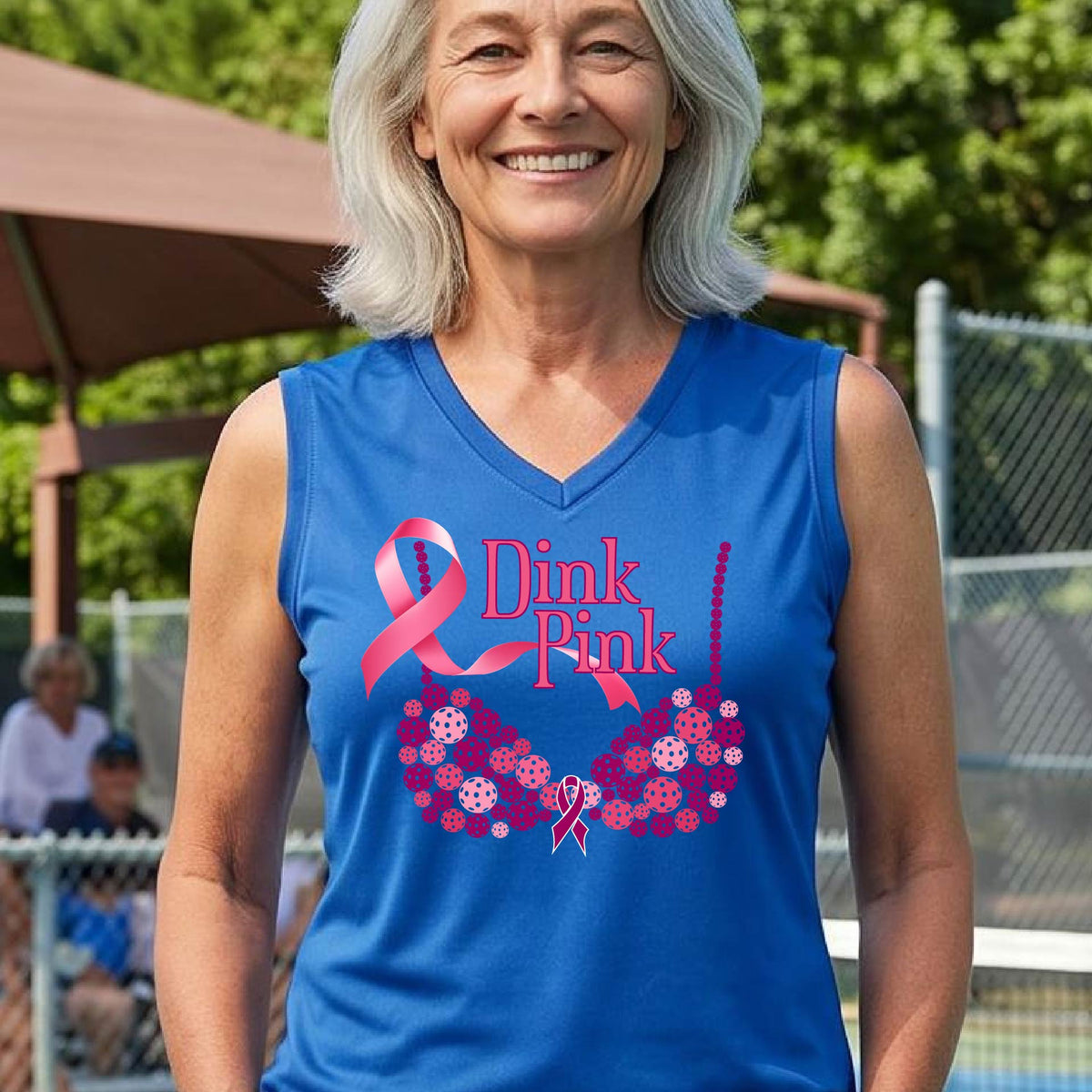A woman on a pickleball court wearing a royal blue sleeveless top with a pink 'Dink Pink' graphic and a pink floral bra design. The background shows a blurred pickleball court and net.