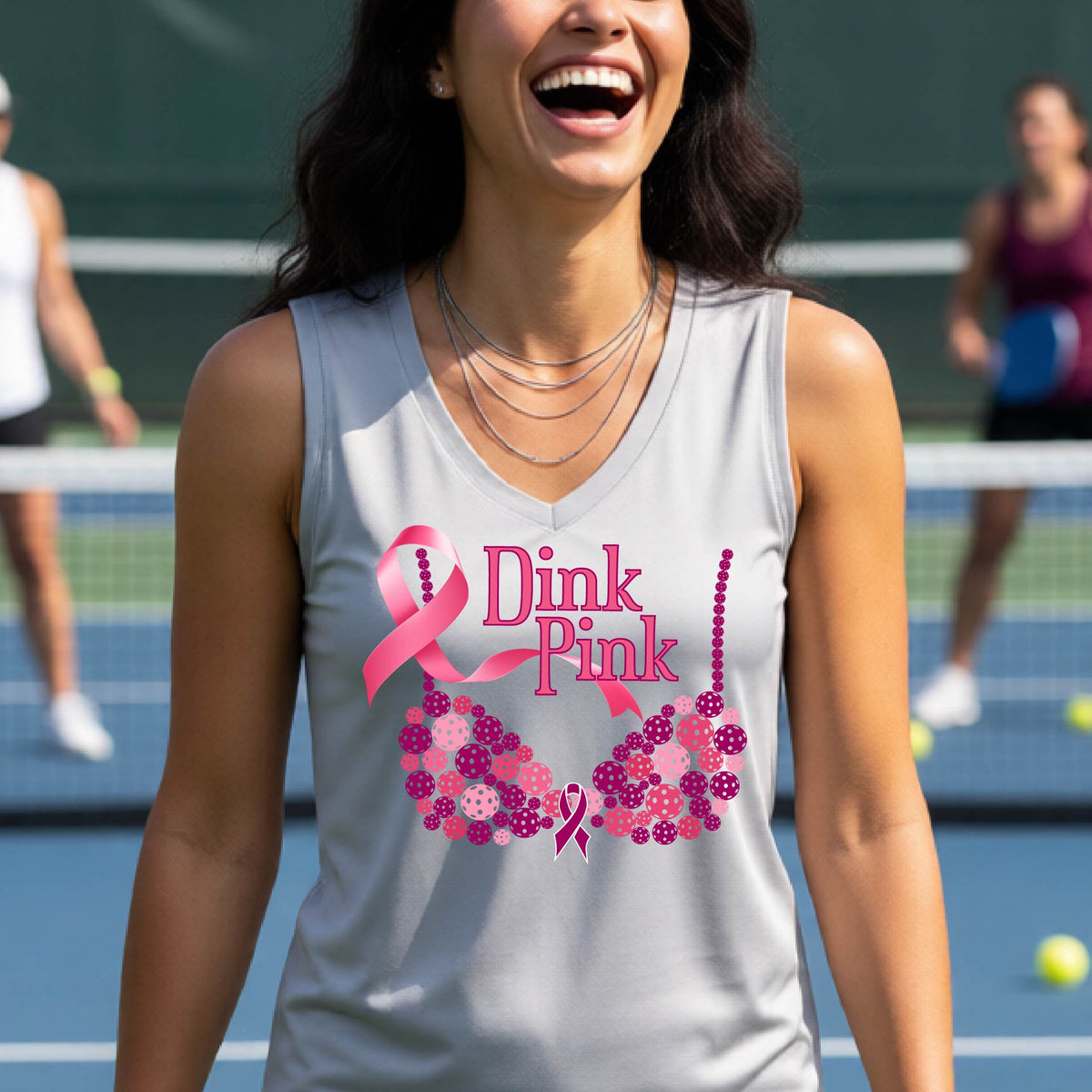A woman on a pickleball court wearing a silver gray sleeveless top with a pink 'Dink Pink' graphic and a pink floral bra design. The background shows a blurred pickleball court and net.